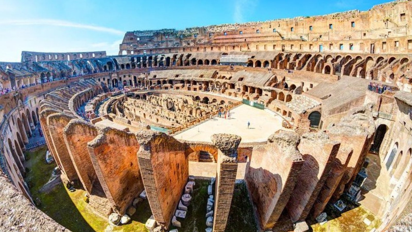 Wide-angle view of the ancient Roman Colosseum ruins in Rome, with its elliptical arena and stone arches under a bright blue sky—an iconic site that belongs on every Italy bucket list, as visitors explore this unforgettable landmark.