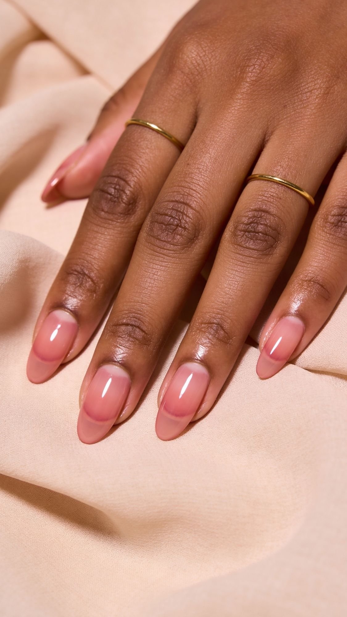 A hand with neatly manicured nails painted in a glossy nude pink shade—perfect for spring nails—rests on soft beige fabric. The person is wearing two thin gold rings on their middle and ring fingers.