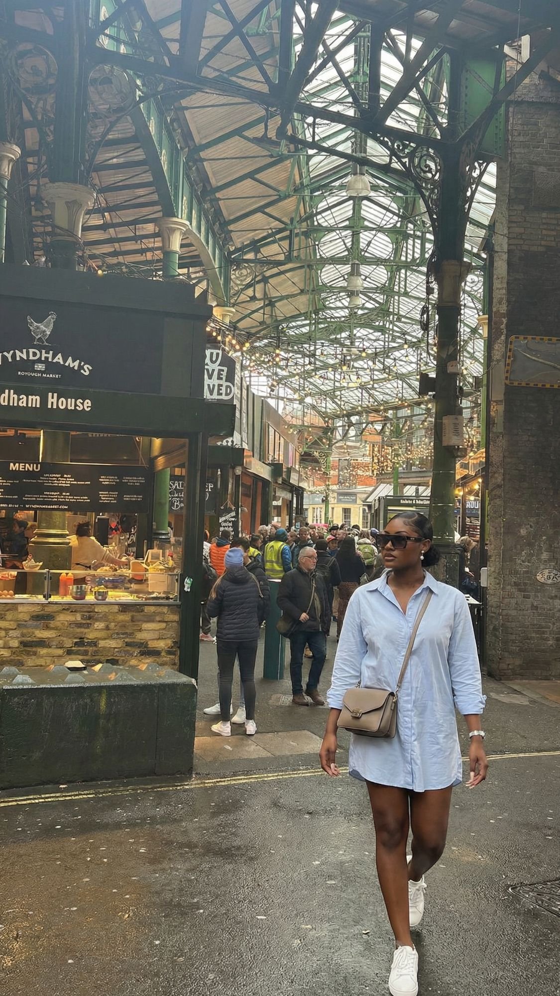 A woman in a light blue shirt dress and sneakers explores a bustling indoor market with glass and metal roofing—a must-see for your first trip to London. Shoppers browse food stalls beneath green metal beams, capturing the essence of London things to do.