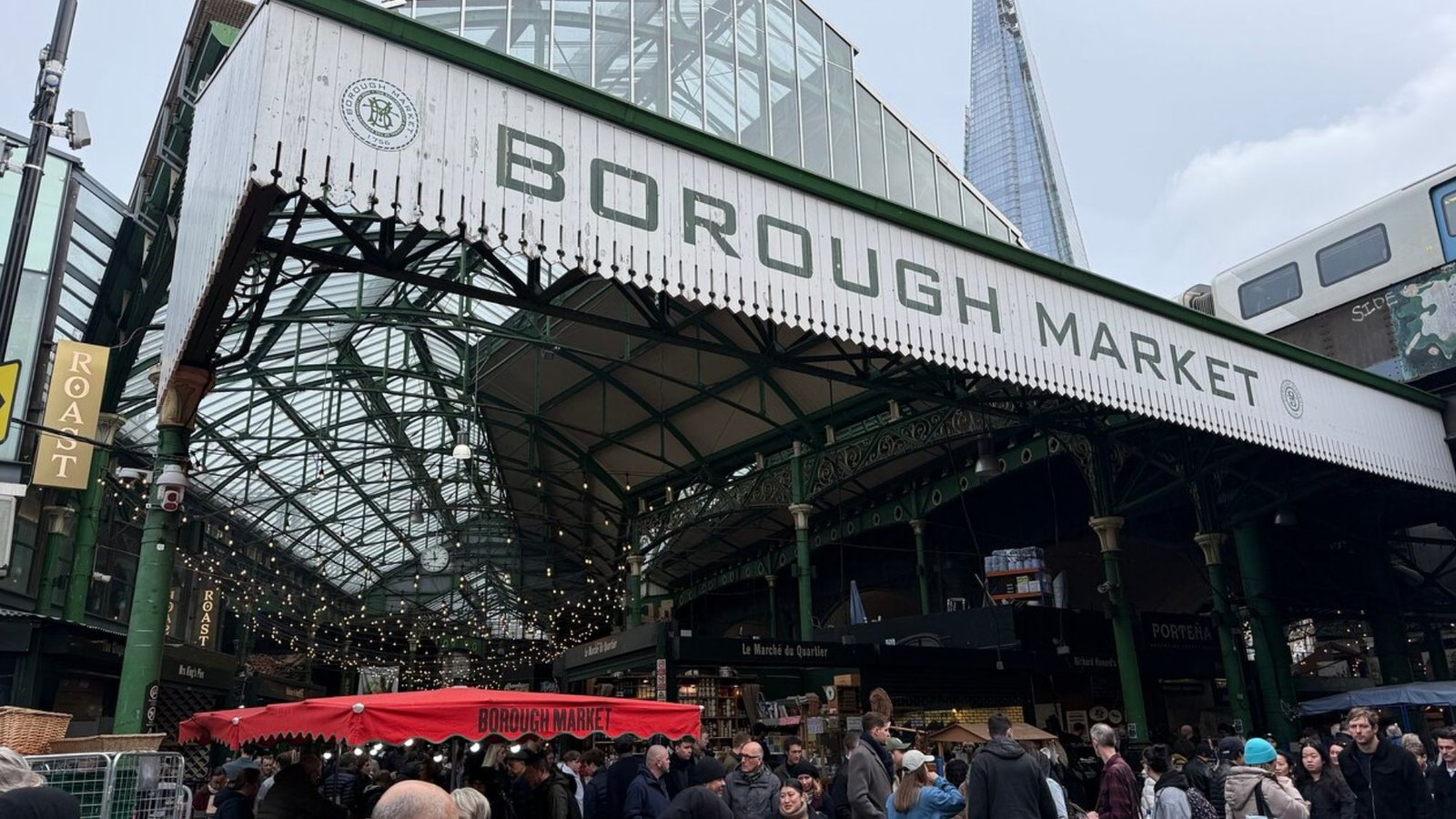 Crowds gather under the large, green entrance of Borough Market in London, with stalls and string lights inside. The Shard rises in the background—a must-see for anyone planning London travel or a first trip to London.