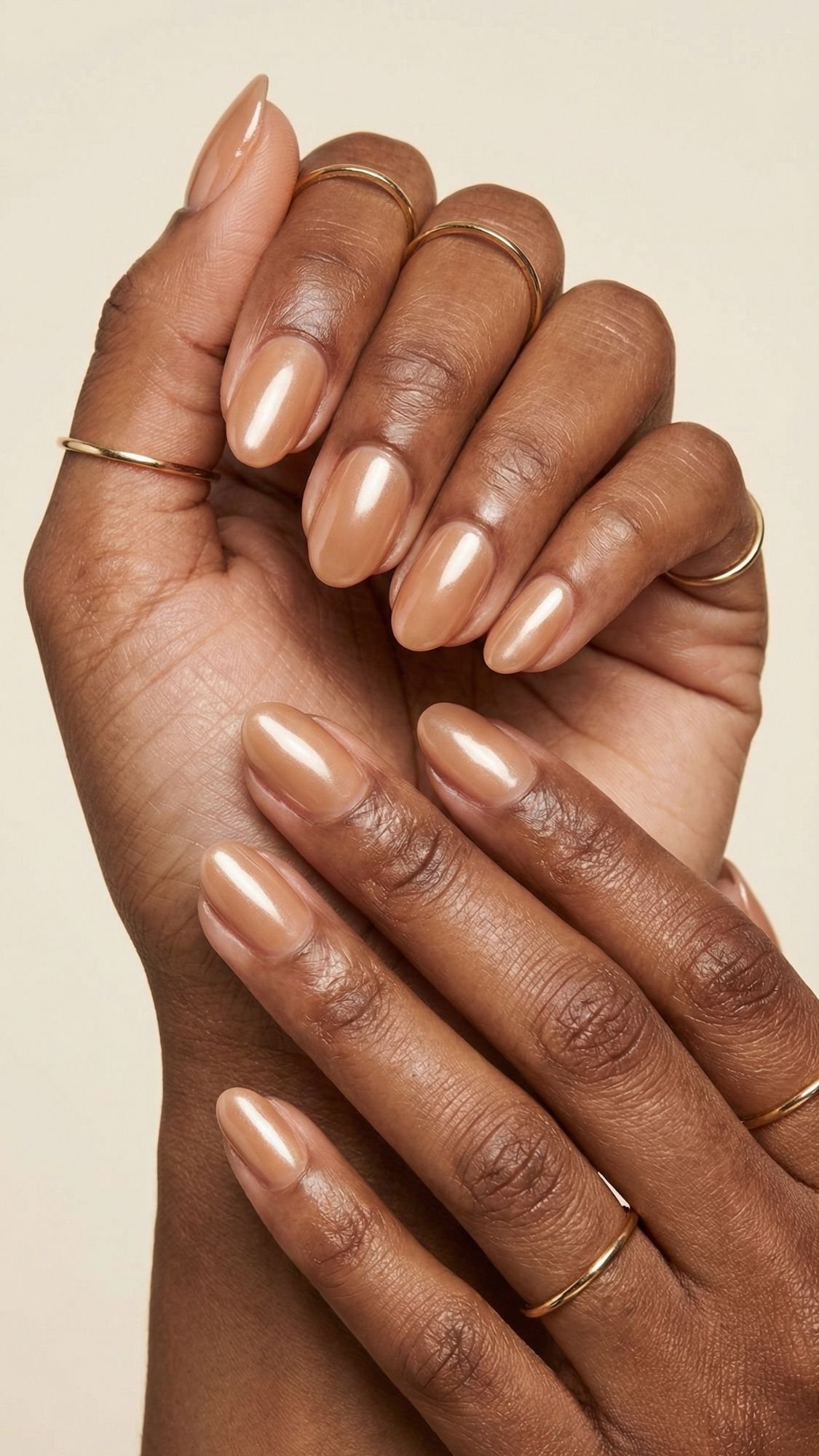 Close-up of two hands with glossy, nude-colored manicured nails—perfect inspiration for nude nail ideas this spring or summer. Fingers are adorned with thin gold rings against a neutral beige background.