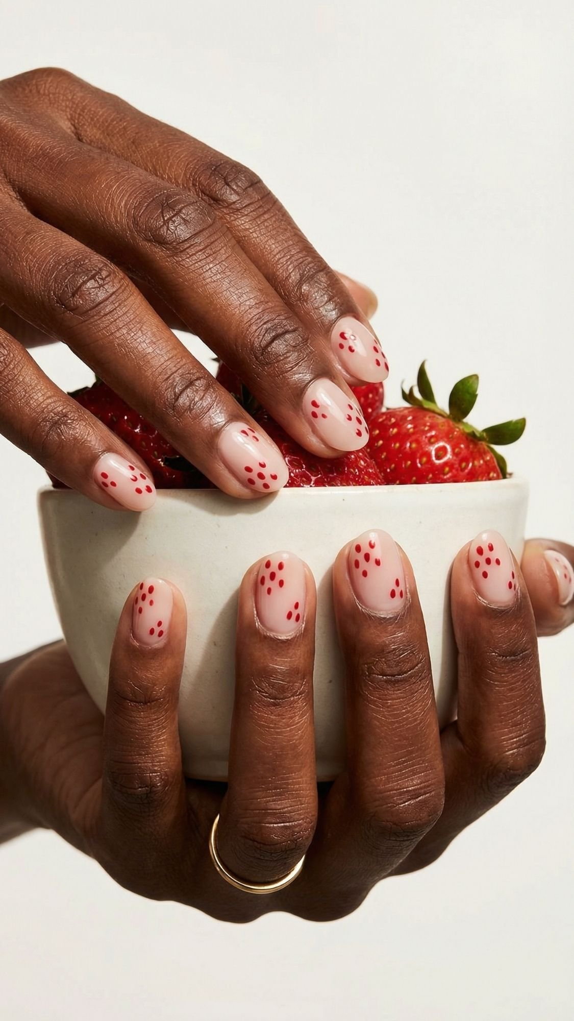 A person with a gold ring holds a white bowl of strawberries. Their nails for every season are painted nude with small red dots, resembling strawberry seeds—a playful nail idea as another hand reaches into the bowl.
