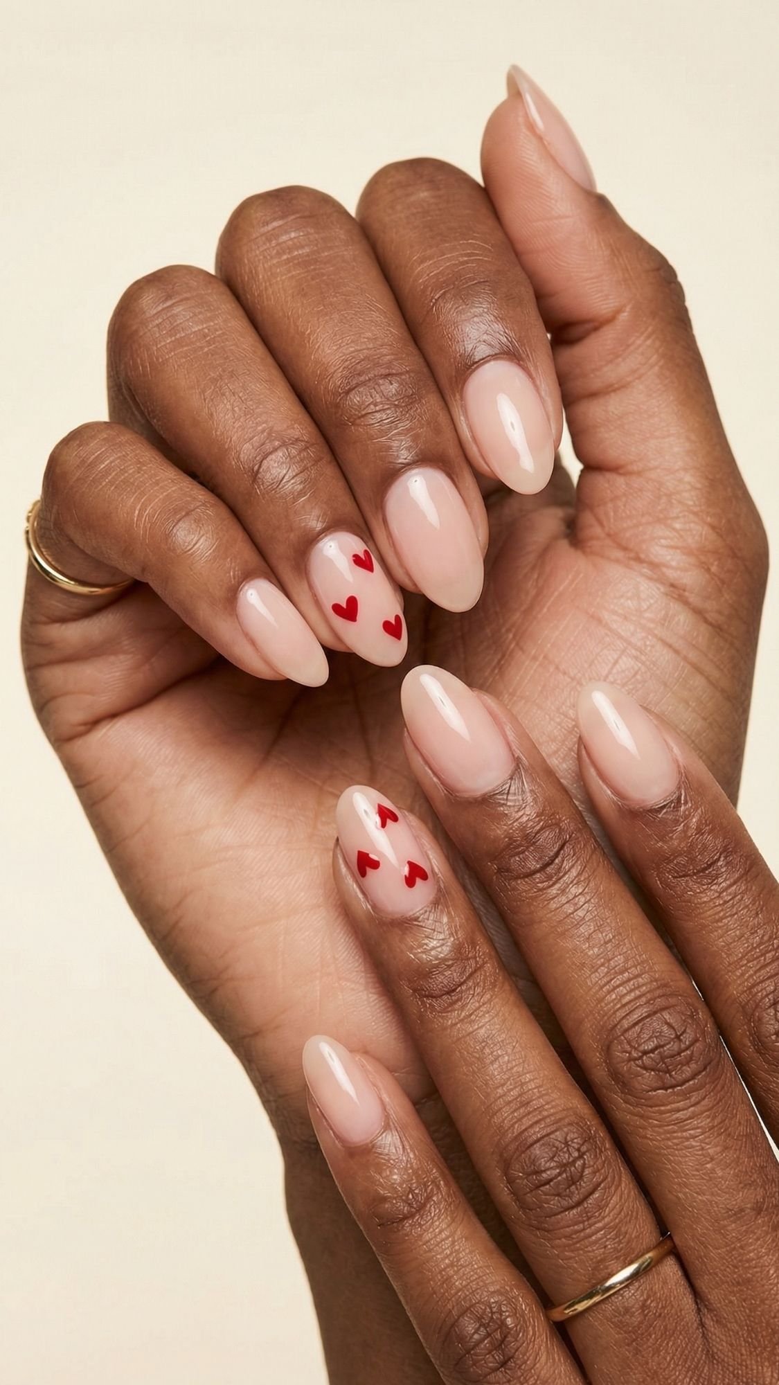 A close-up of hands with almond-shaped nude nails showcases adorable red heart designs—perfect nail ideas for seasonal nails. The person is wearing thin gold rings on two fingers.