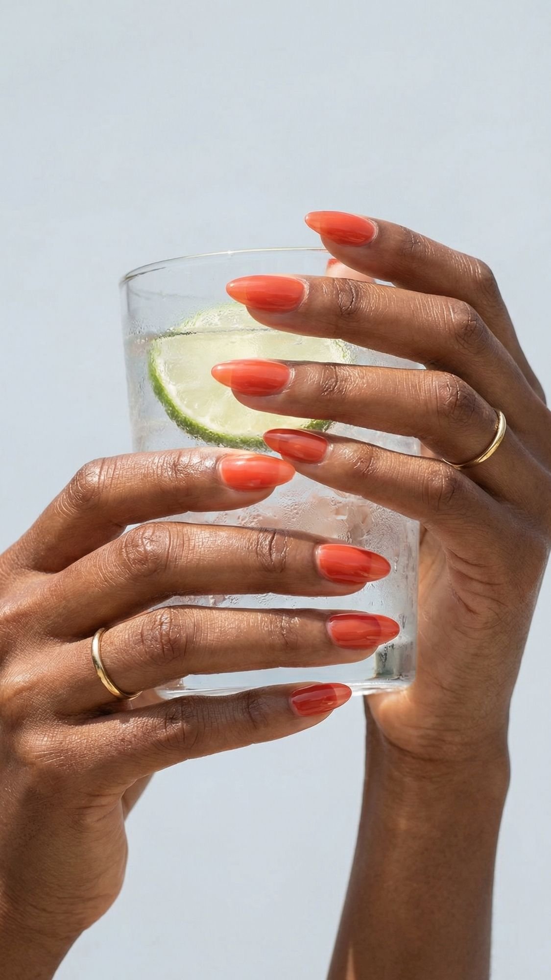 Two hands with red nails and gold rings hold a clear glass of sparkling water with a slice of lime against a light background—fresh nail ideas perfect for every season.