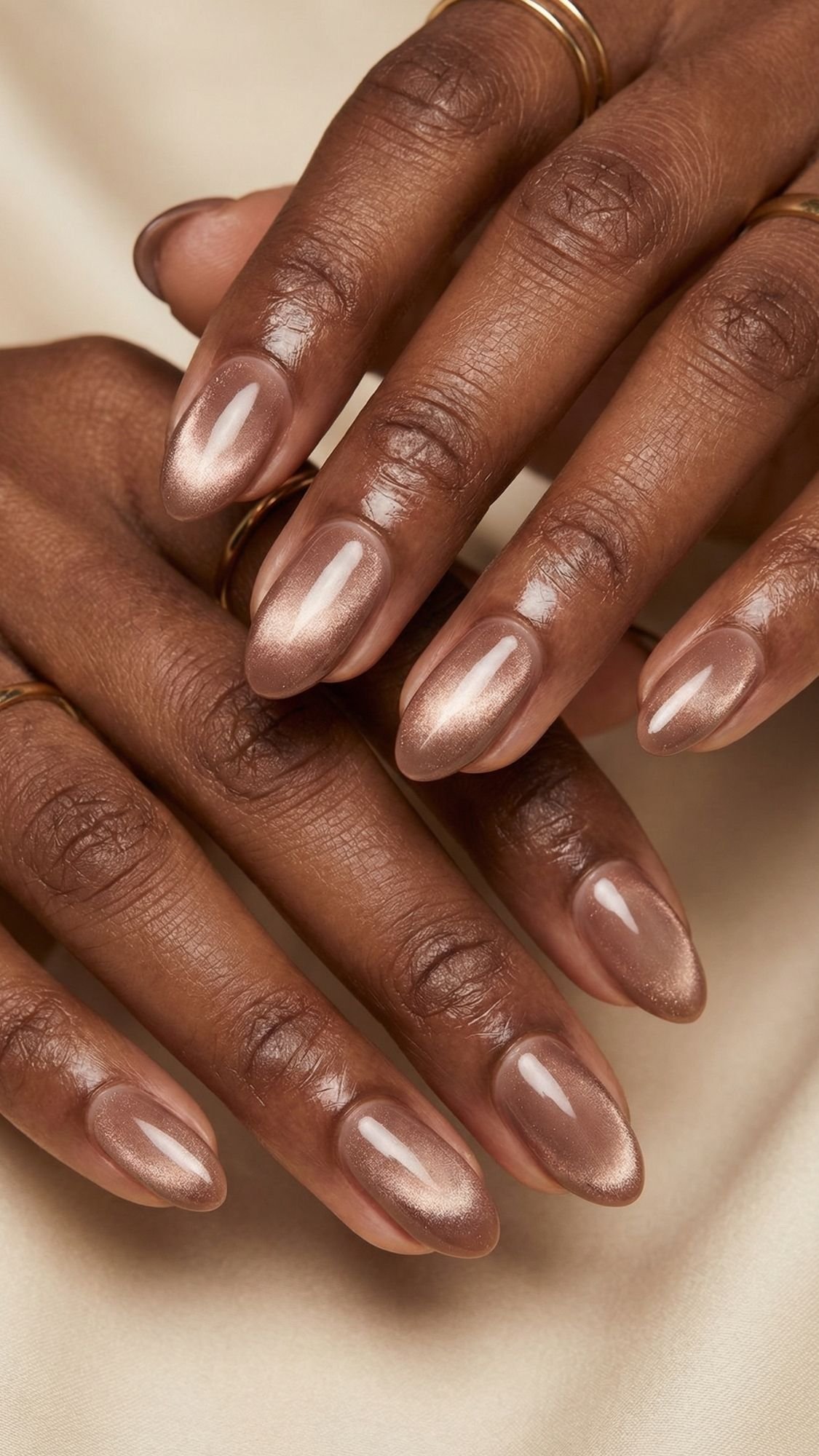 Close-up of hands with medium brown skin, wearing thin gold rings and featuring almond-shaped nails in shimmery nude polish—perfect inspiration for spring nails—resting on a beige satin fabric background.