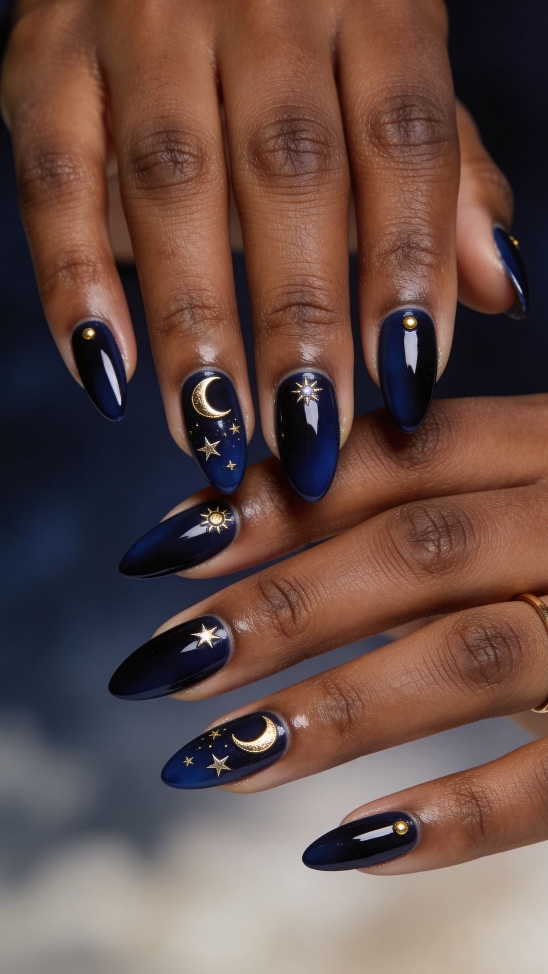 Close-up of hands with long, almond-shaped nails painted dark blue and adorned with gold crescent moons, stars, and studs—a celestial look perfect for quick nail designs you can try on simple nails at home. A thin gold ring adds an elegant touch.