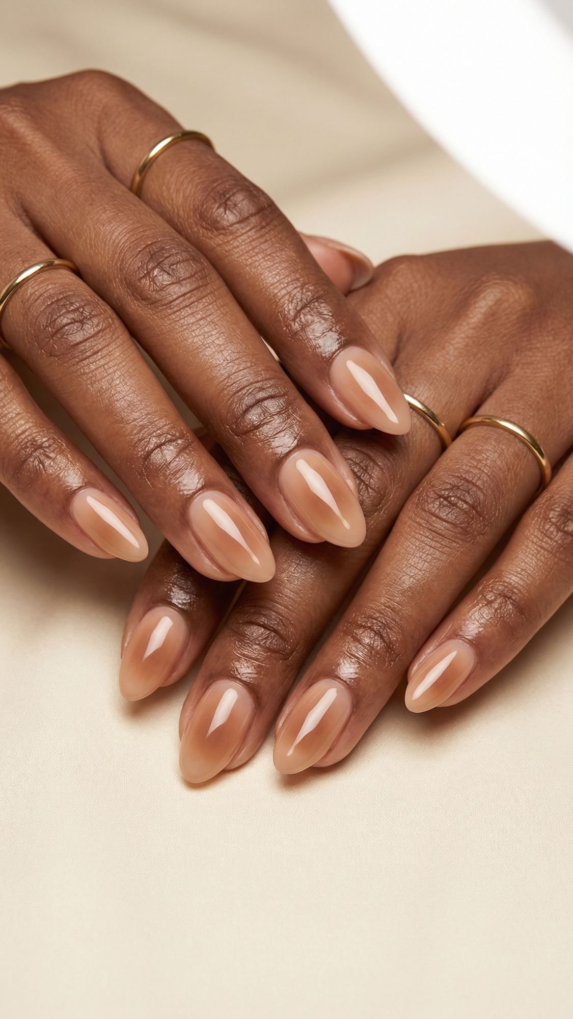 Close-up of well-groomed hands with almond-shaped nude glossy nails, a perfect choice for spring nails, wearing thin gold rings and resting on a light beige surface.
