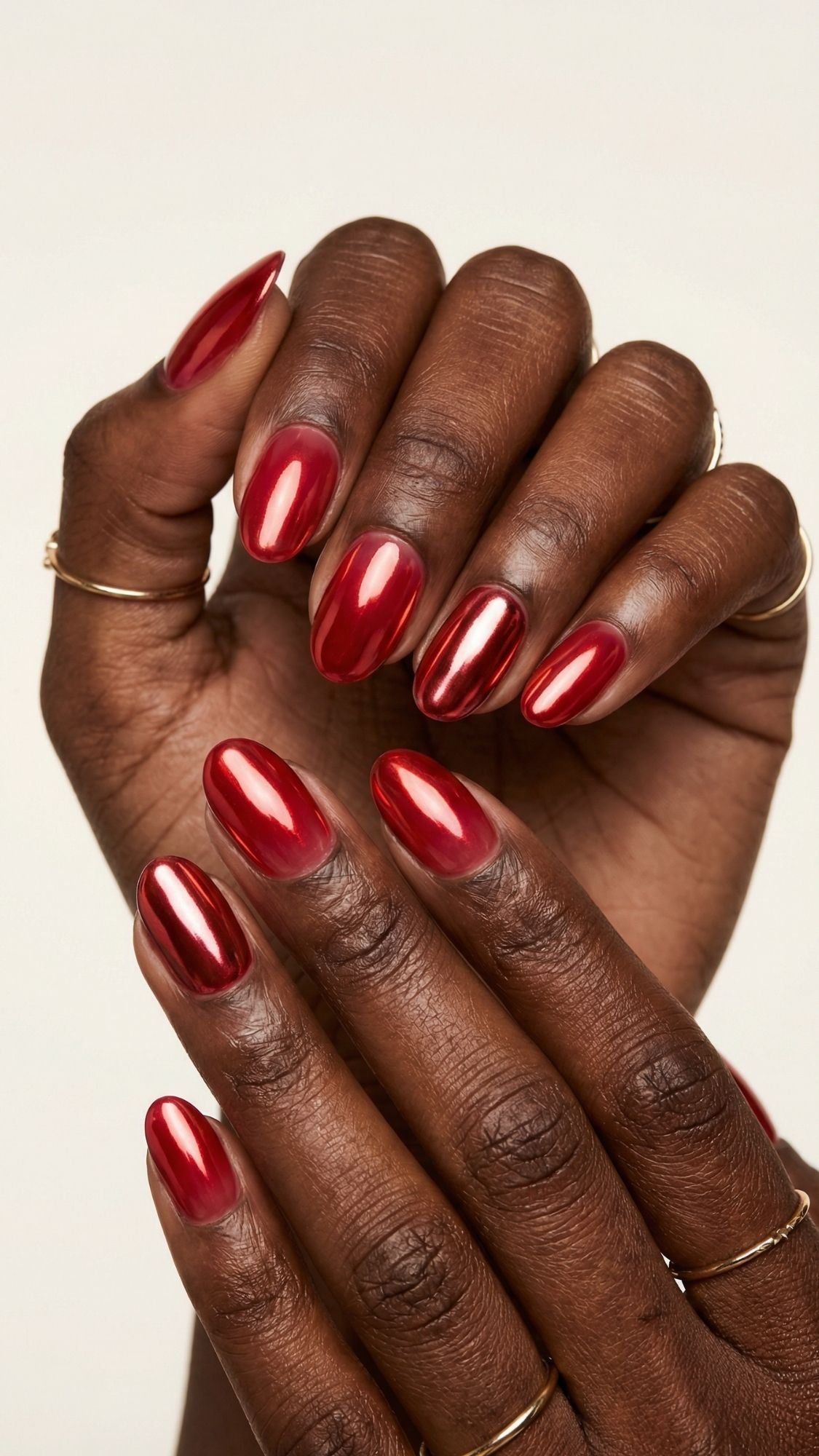 A close-up of hands with dark skin tone, featuring shiny, red almond-shaped nail polish—an elegant red nails look perfect for every season. Thin gold rings adorn the fingers, which are posed gracefully against a neutral background.