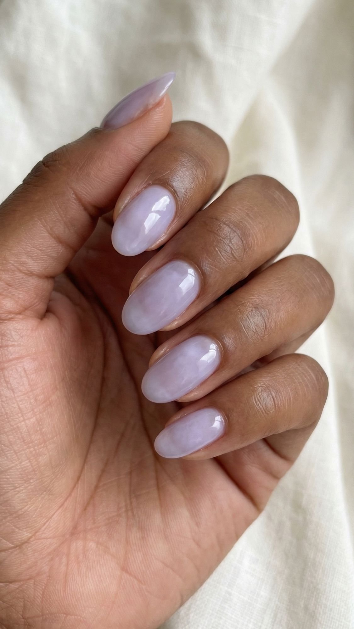 A hand with medium-toned skin shows almond-shaped nails painted with a glossy, pale lavender polish—perfect for simple nails that you can recreate easily at home—against a soft, light fabric background.