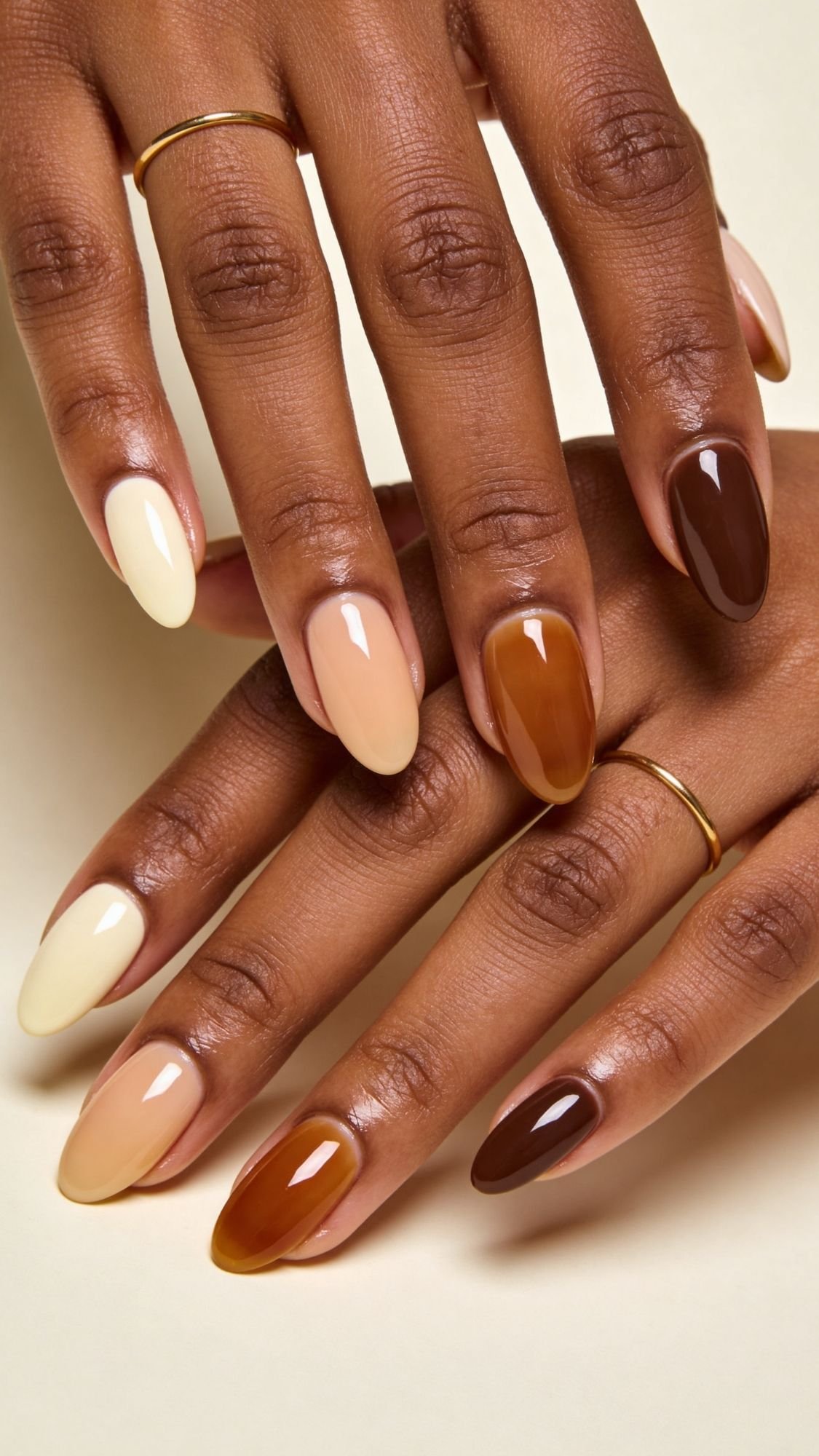 A close-up of hands with almond-shaped nails painted in different shades of beige, brown, and cream—perfect nude nail ideas for spring nails. Simple gold rings adorn the fingers against a light neutral background.
