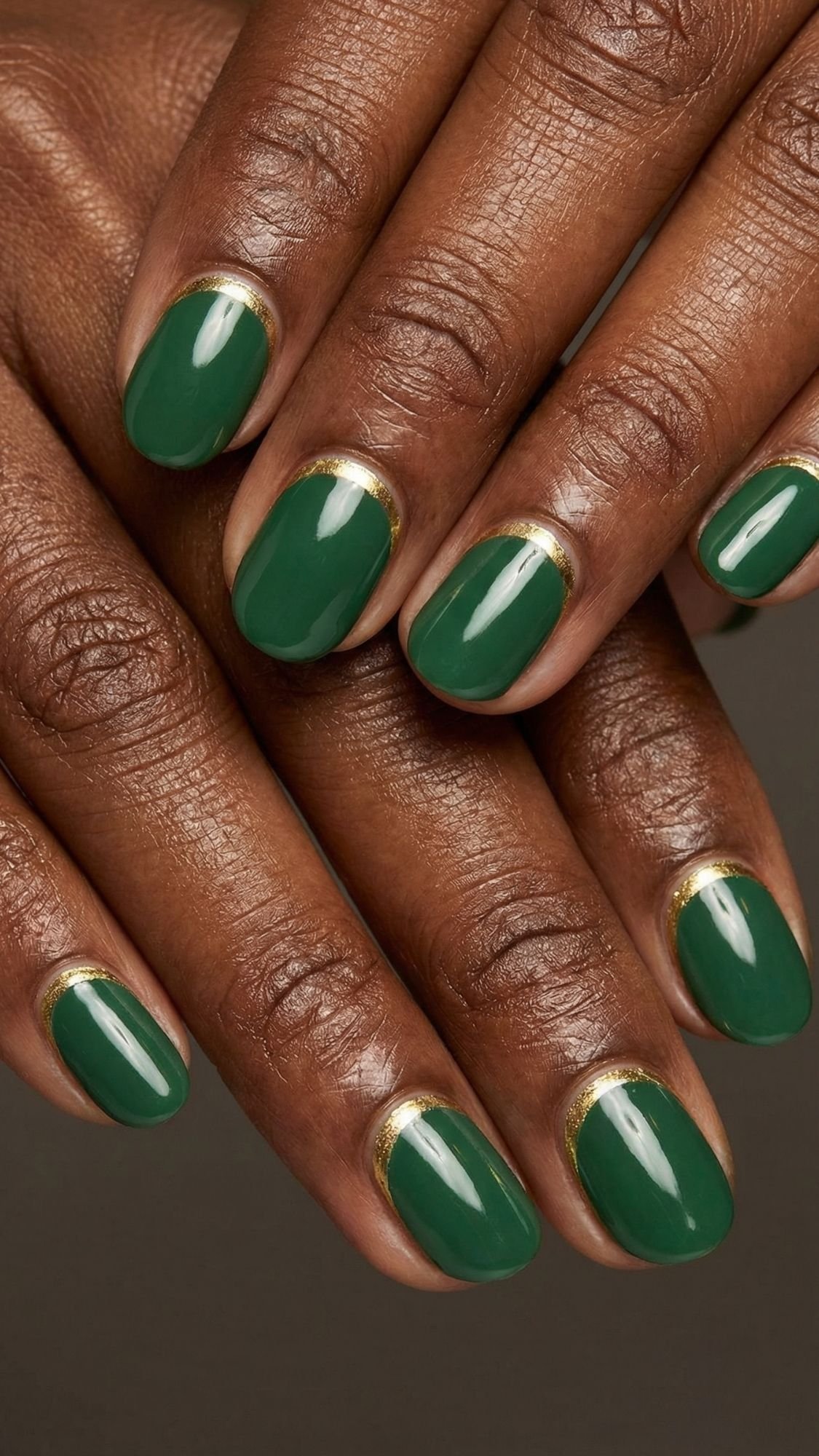 A close-up of hands with neatly manicured, simple nails painted green, featuring a thin gold accent line along the base. The skin tone is deep brown, and the short, rounded nails make this an easy nail design to try for nails at home.