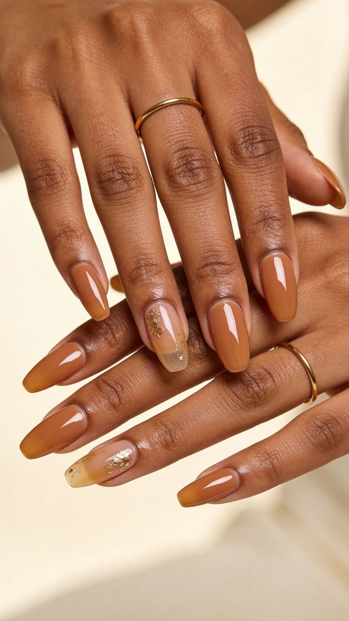 Close-up of hands with almond-shaped nails painted in a glossy caramel brown shade—perfect for spring nails. Accent nails feature clear tips with gold foil details, complemented by simple gold rings on several fingers.