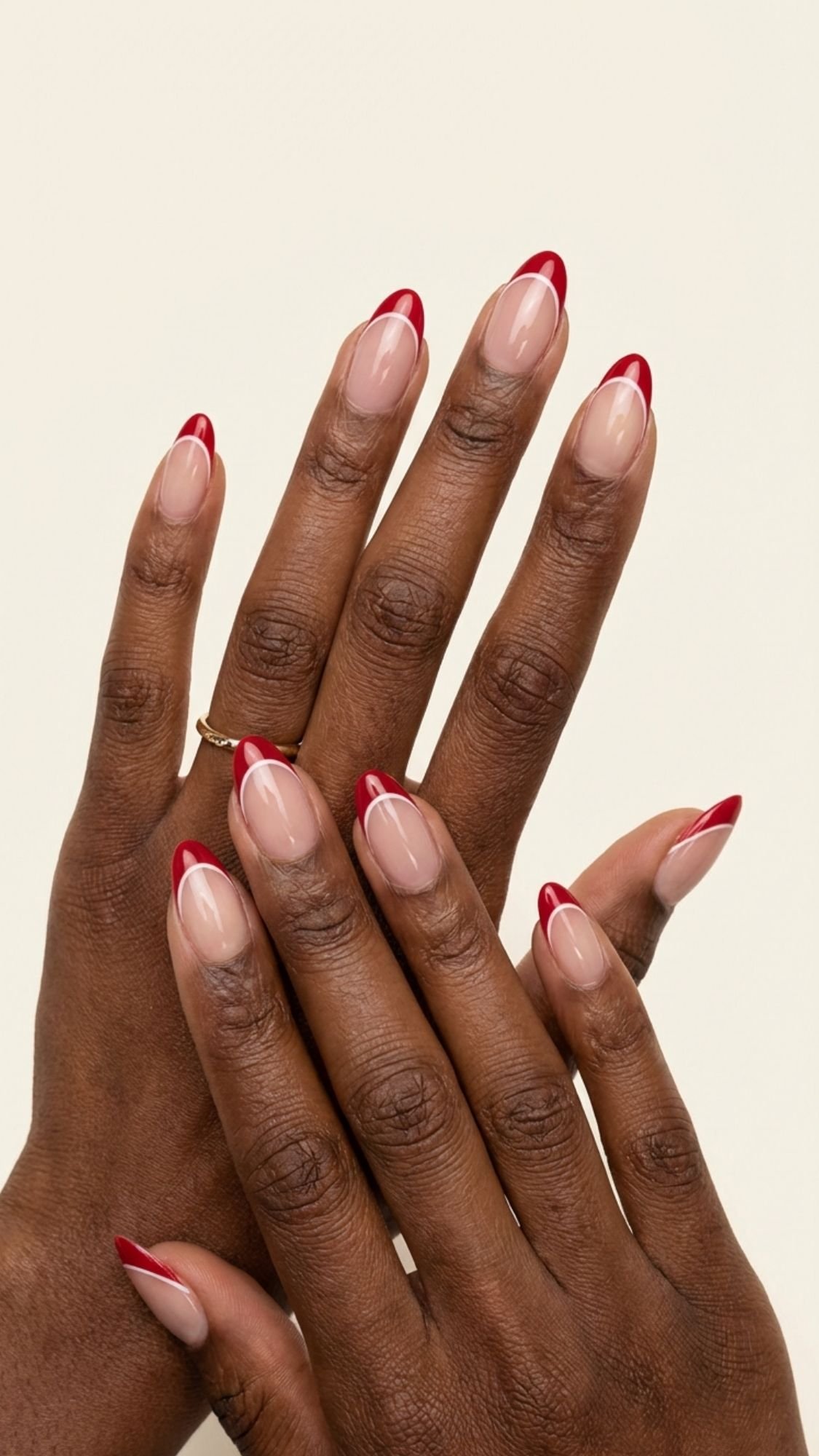 Two hands with medium-brown skin display almond-shaped nails featuring a nude base and bold red French tips—perfect for those seeking fresh nail ideas. One hand wears a simple gold ring, set against an off-white background.