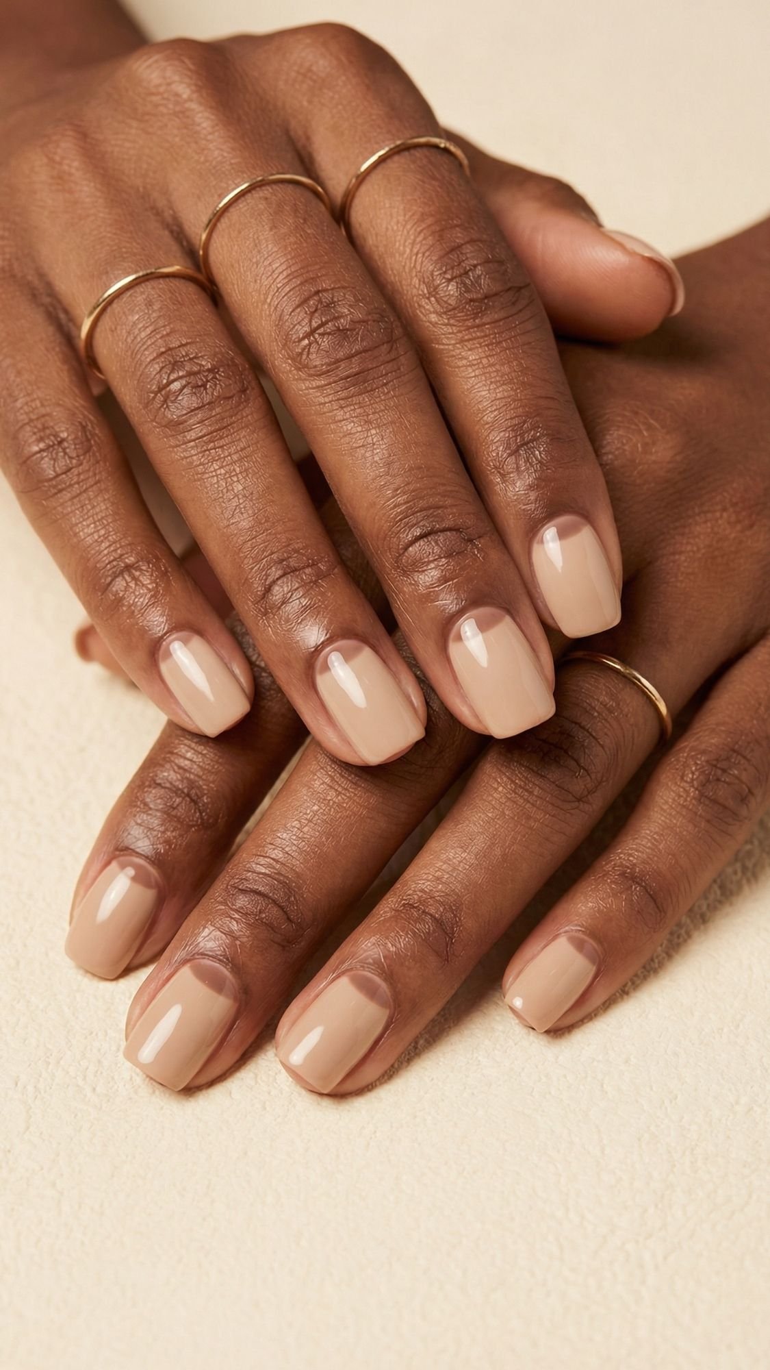 Close-up of hands with neatly manicured summer nails in a glossy nude shade, wearing thin gold rings on several fingers, resting on a light textured surface.