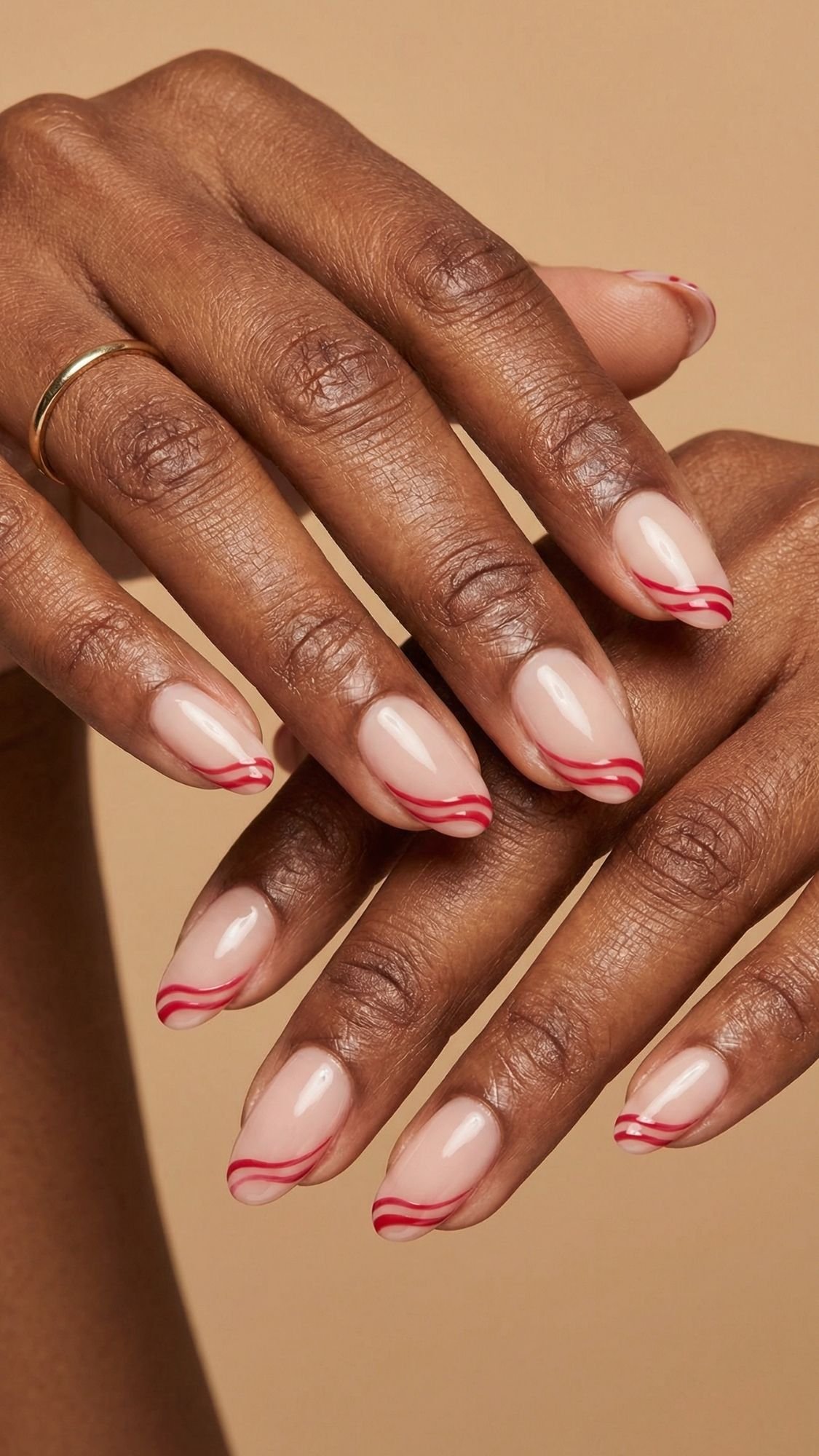 A close-up of hands with almond-shaped nails painted in a nude color and decorated with thin, wavy red lines along the tips—chic nail ideas for every season. One hand wears a simple gold ring on the ring finger. The background is beige.