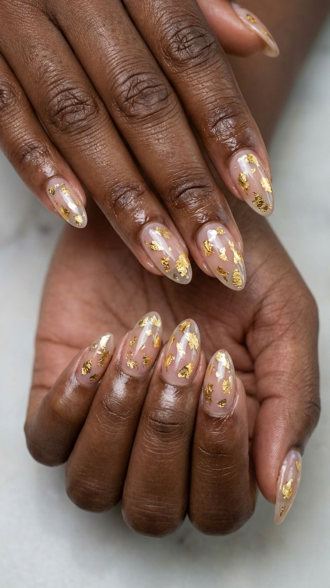 Close-up of hands with almond-shaped nails featuring a clear nude base and gold foil accents, creating elegant and modern nail art. Perfect for simple nails you can achieve as easy nail designs for stunning nails at home. Skin tone is deep and the nails are well-manicured.