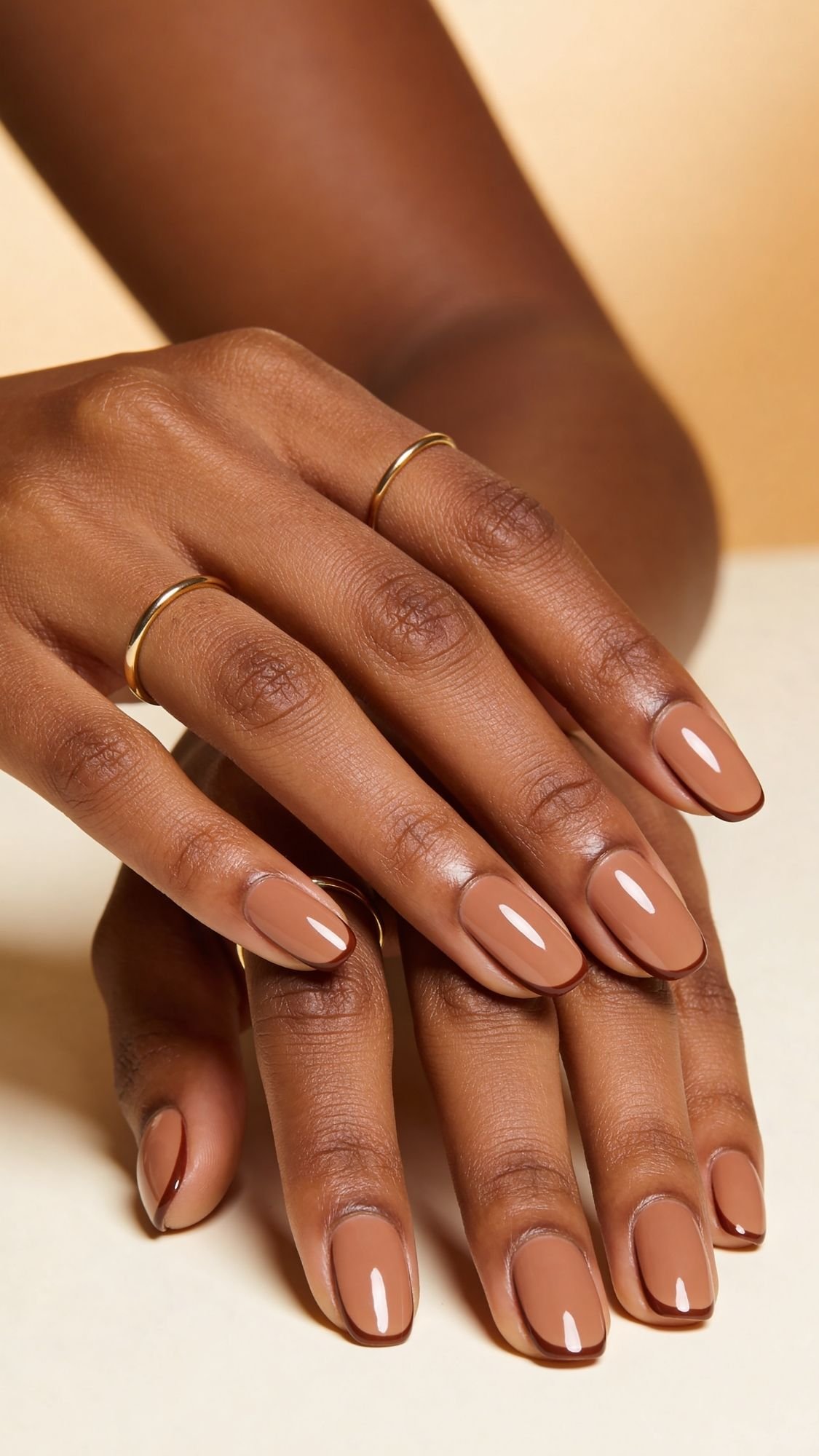 Close-up of hands with medium brown glossy nail polish, perfect for summer nails, and two thin gold rings on the middle and ring fingers, against a neutral beige background.