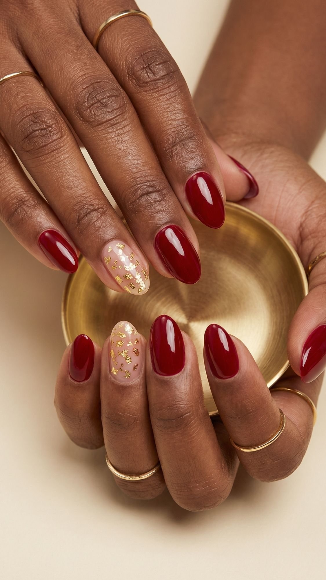 A close-up of hands with oval-shaped red nails, perfect for seasonal nails inspo, except for one nude accent nail with gold glitter. The hands hold a small, round gold-colored dish and are adorned with thin gold rings.