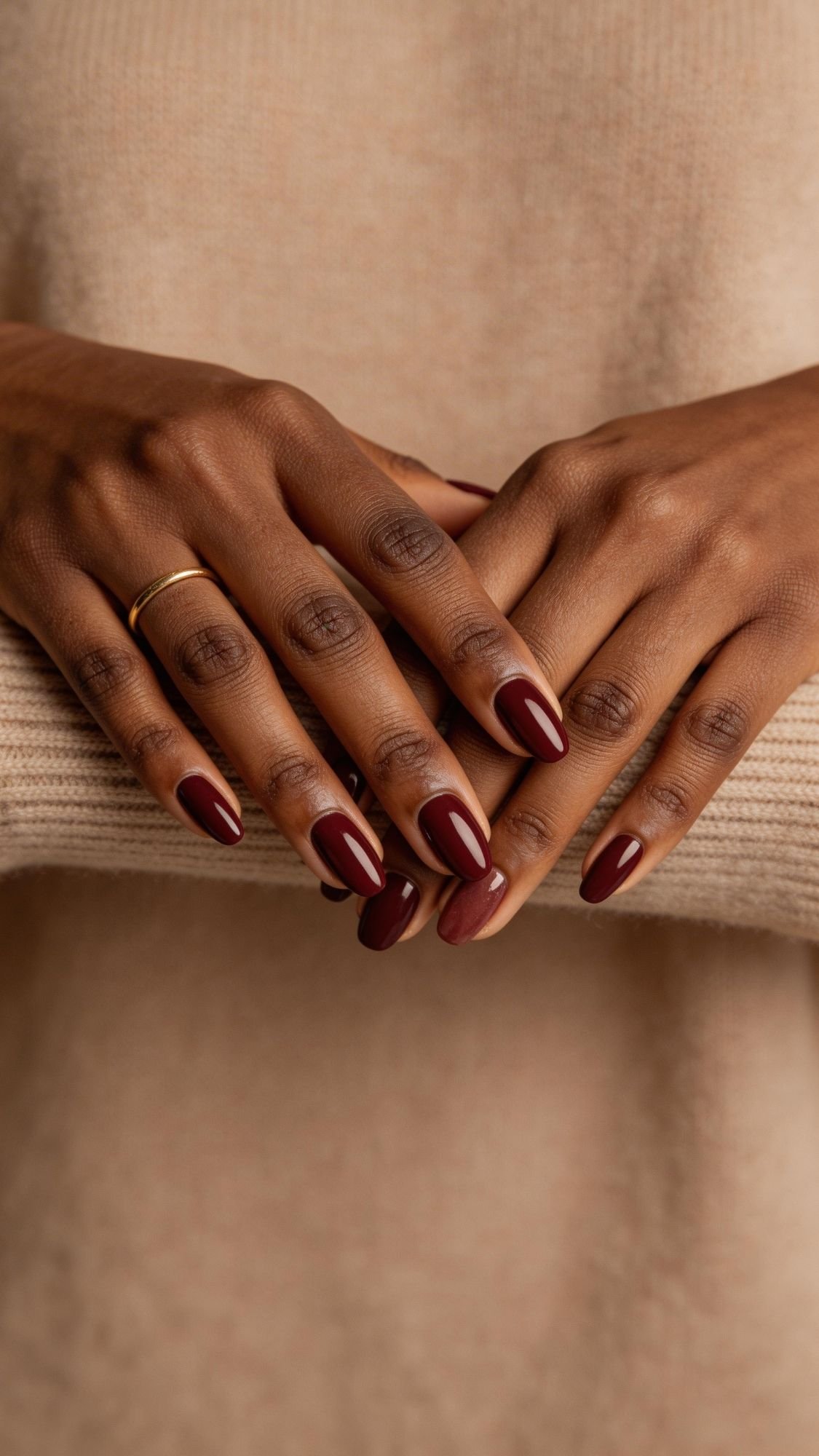 Close-up of two hands resting on a beige knit sweater, showing neatly manicured red nails in a glossy dark burgundy shade—chic nail ideas perfect for every season. One hand has a simple gold ring on the ring finger.