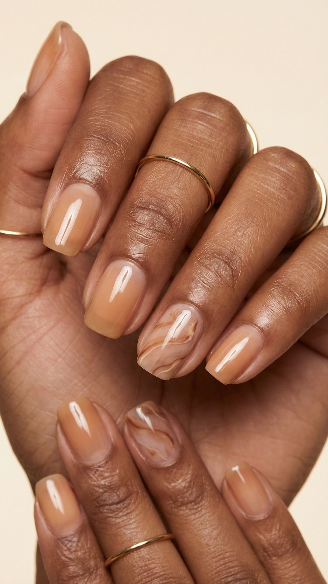 A close-up of hands with neatly manicured nails painted in a glossy nude shade, featuring subtle marble designs on two accent nails—perfect nude nail ideas for chic summer nails. Thin gold rings adorn a few fingers for added elegance.