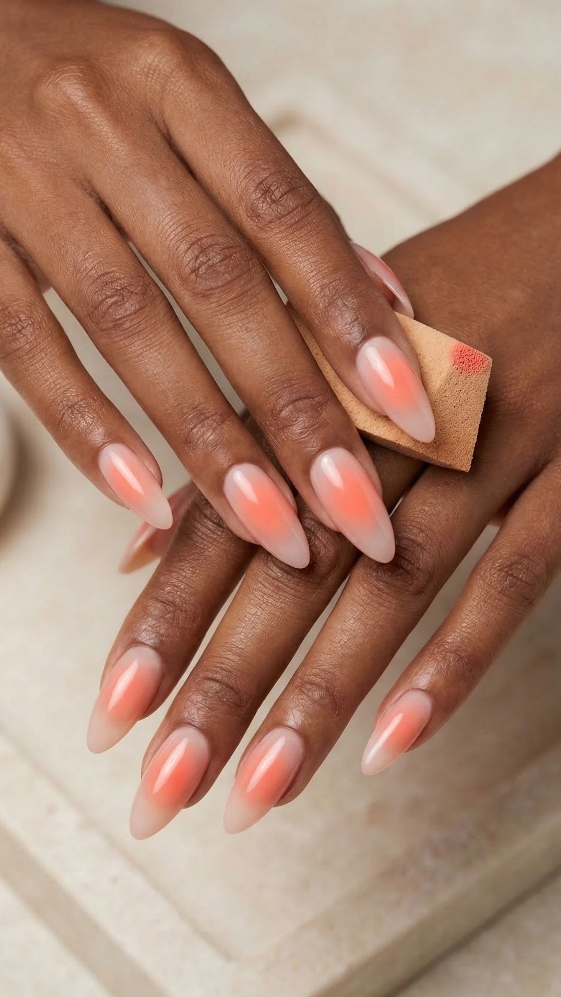 A close-up of hands with almond-shaped nails featuring a glossy, gradient coral and nude manicure—perfect inspiration for simple nails you can achieve with DIY nails techniques at home. The person holds a small beige makeup sponge with a coral lipstick mark on it.