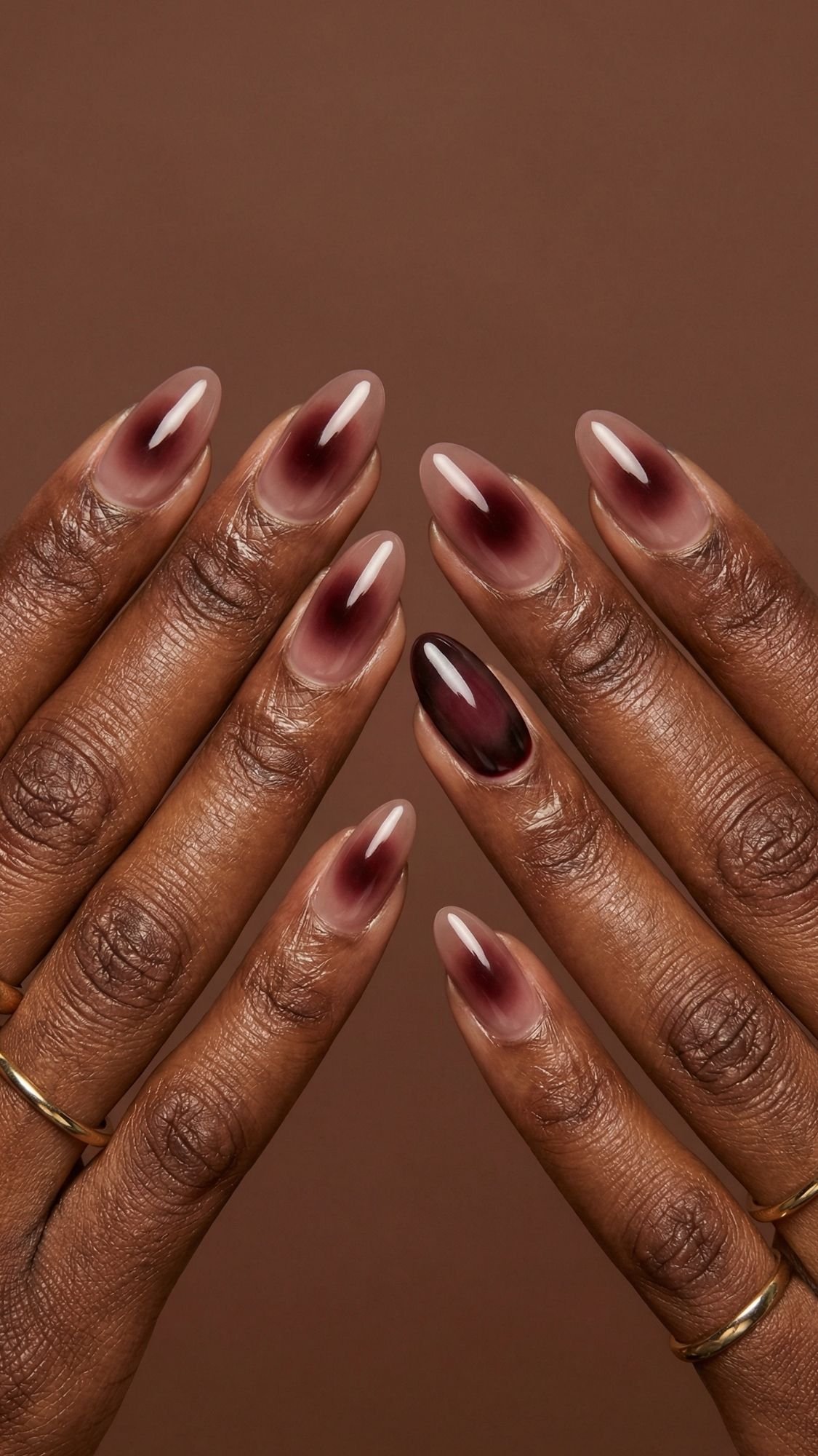Two hands with almond-shaped nails featuring a gradient ombré design in deep burgundy and nude tones, perfect for seasonal nails inspiration. The person wears simple gold rings, shown against a brown background.