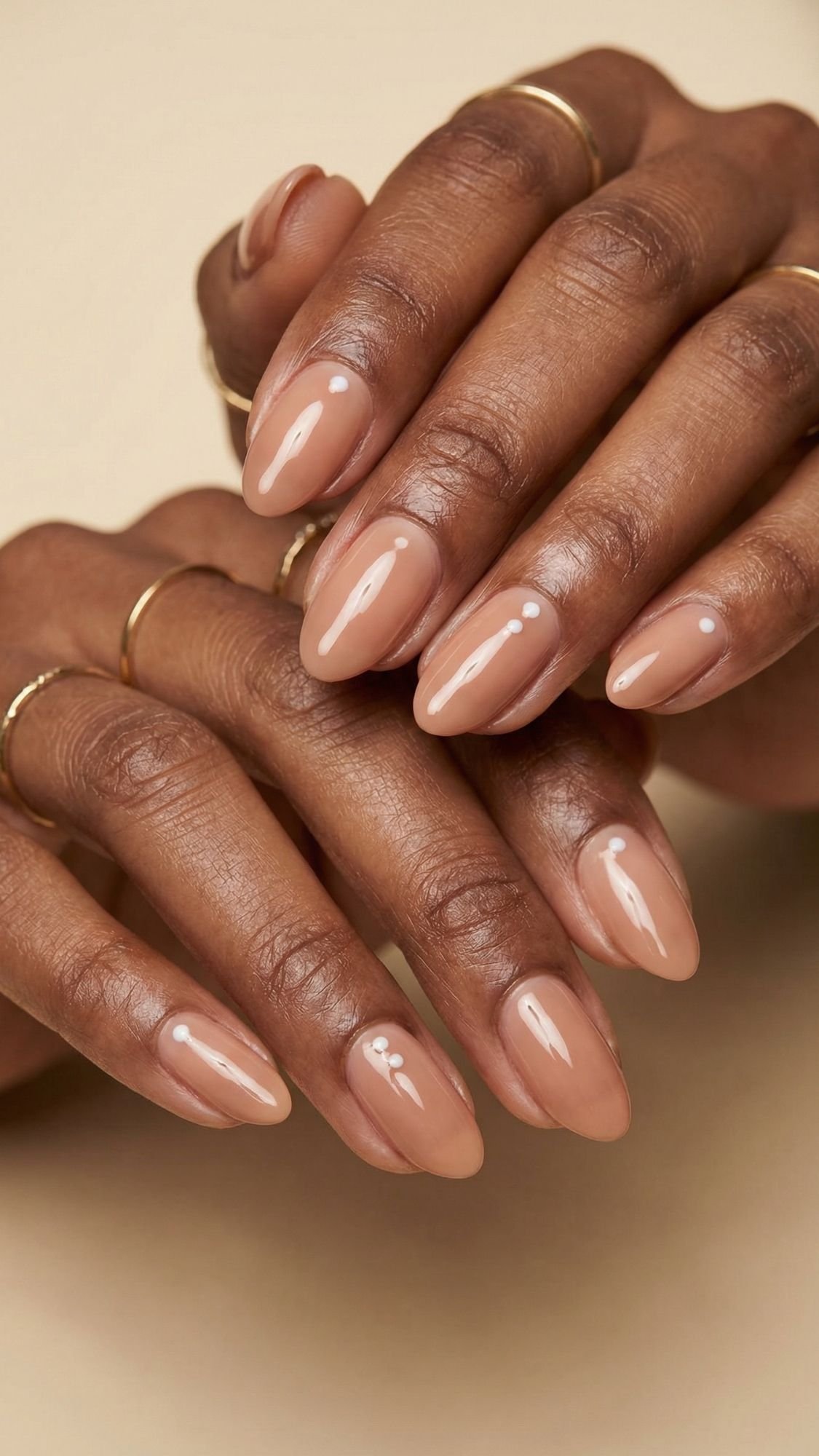 Two hands with almond-shaped, nude-colored manicured nails featuring a glossy finish and tiny white dot accents—perfect for nude nail ideas or effortless summer nails. Several thin gold rings adorn the fingers against a beige background.