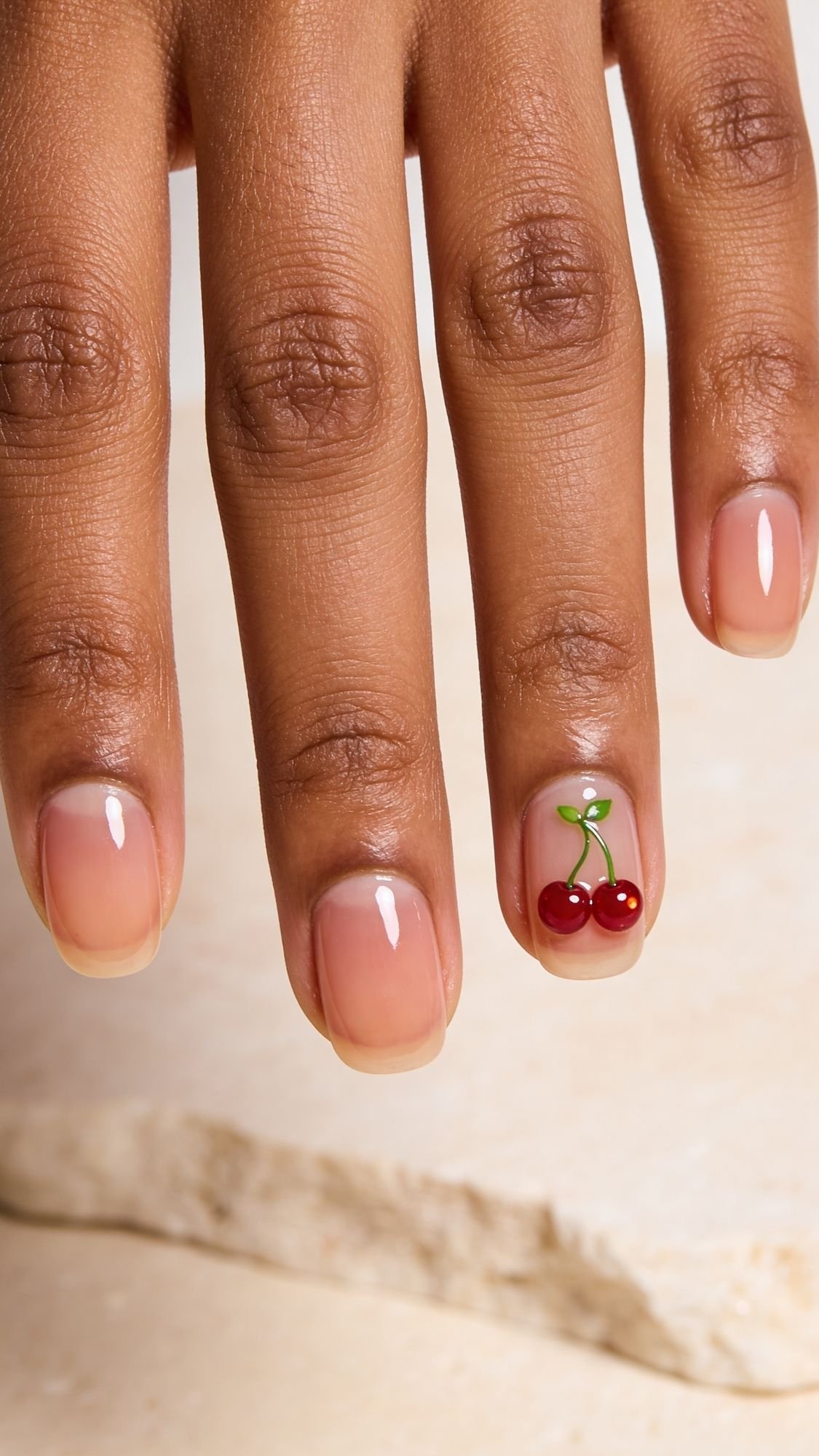 A hand with neatly manicured nails features a simple cherry nail art design on the pinky finger—two red cherries with green stems on a natural, glossy base. Perfect for those looking for easy nail designs to try on their nails at home.