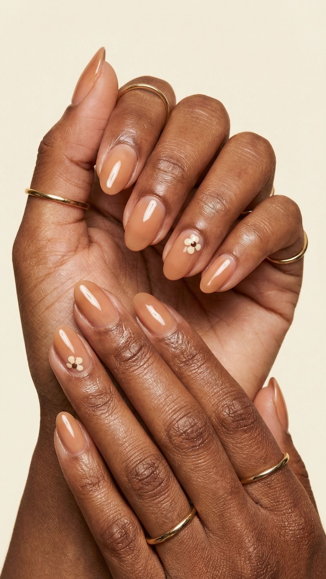Two hands with almond-shaped, glossy nude nails—perfect for fresh spring nails. The ring fingernail on each hand features a small white floral design with a gold center. Thin gold rings add elegance against a beige background.