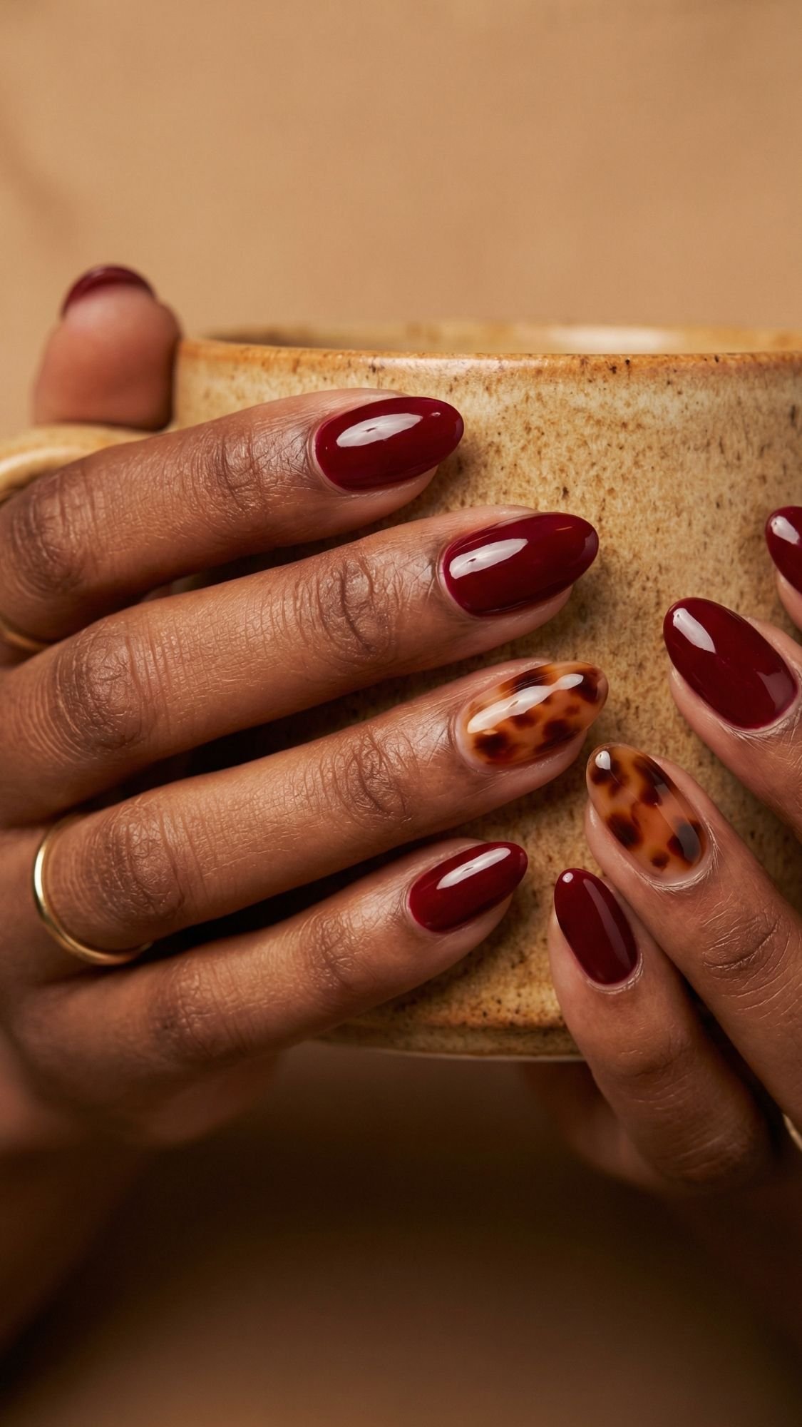 Close-up of hands holding a ceramic mug, showcasing chic red nails with a tortoiseshell accent—an elegant nail idea perfect for every season. A simple gold ring adds a touch of understated style.
