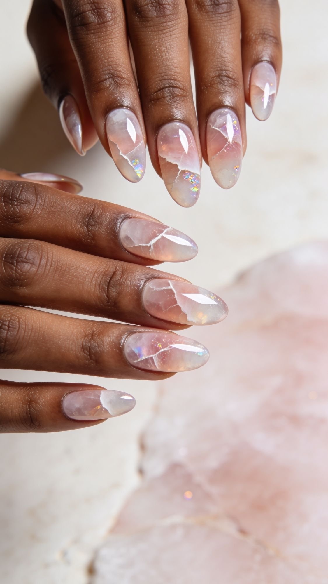 A close-up of hands with almond-shaped nails featuring a translucent, marble-like design in soft pink and white tones, with subtle gold and iridescent accents. These simple nails have a glossy finish and resemble polished gemstones.