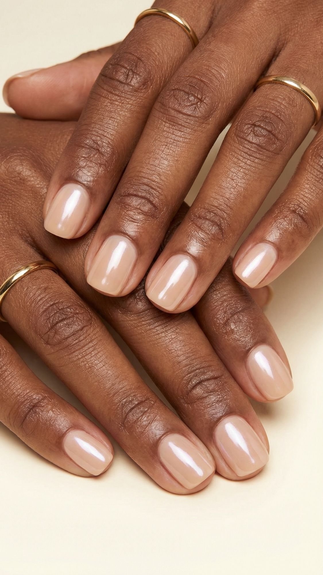 Close-up of hands with short, neatly manicured nails painted in a glossy nude polish—perfect for spring nails. The person wears simple gold rings, set against a soft cream background.
