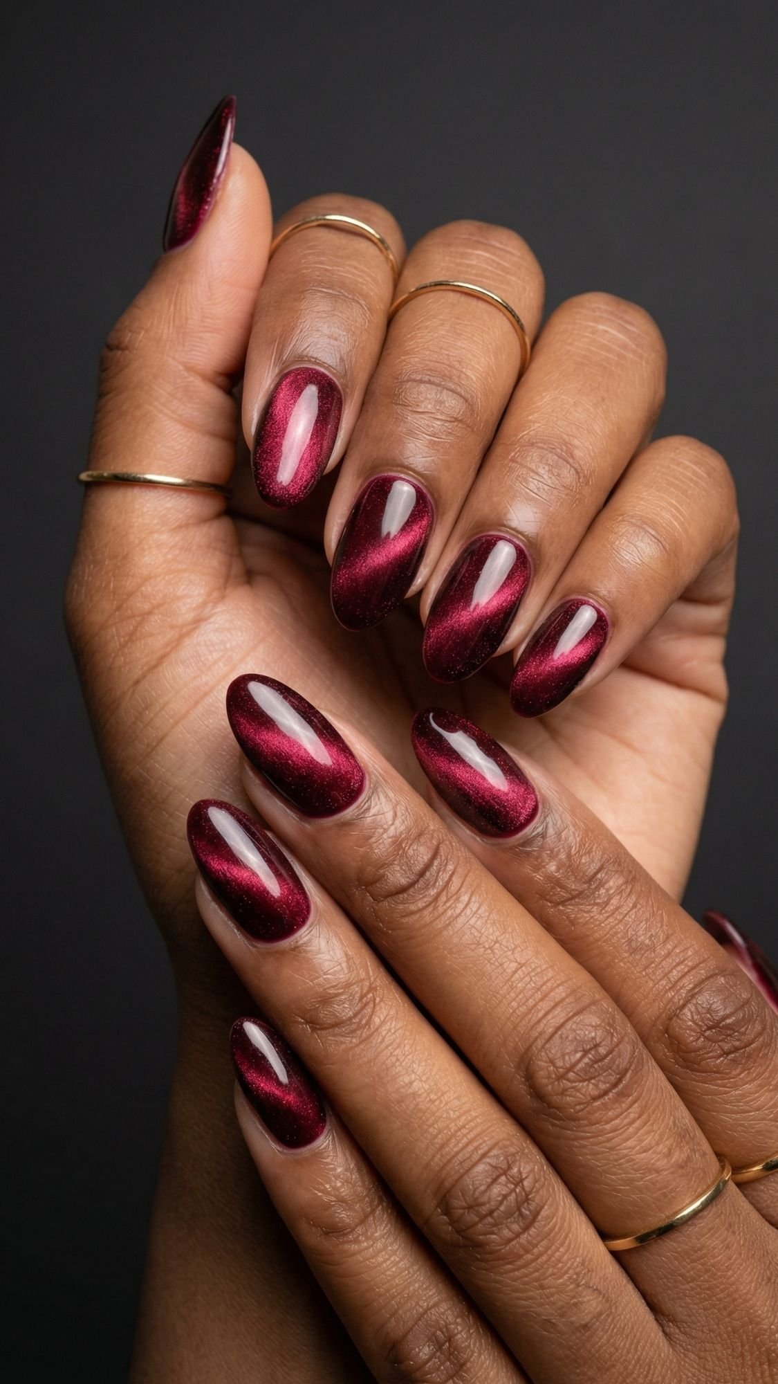 Close-up of hands with dark skin tone, adorned with thin gold rings and glossy, dark red oval-shaped manicured nails—an elegant look for anyone seeking red nail ideas—posed against a black background.