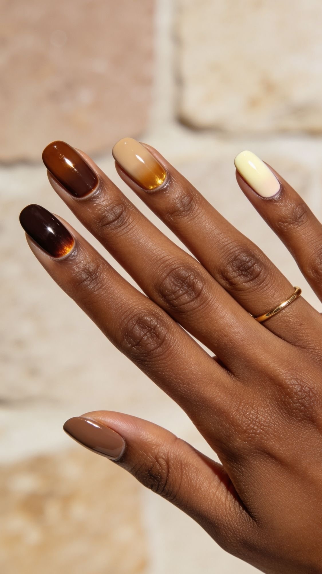 A hand with a gold ring displays simple nails featuring long, almond-shaped tips painted in gradient shades of brown, gold, and cream—an inspiring look for easy nail designs to try with nails at home against a blurred beige stone background.