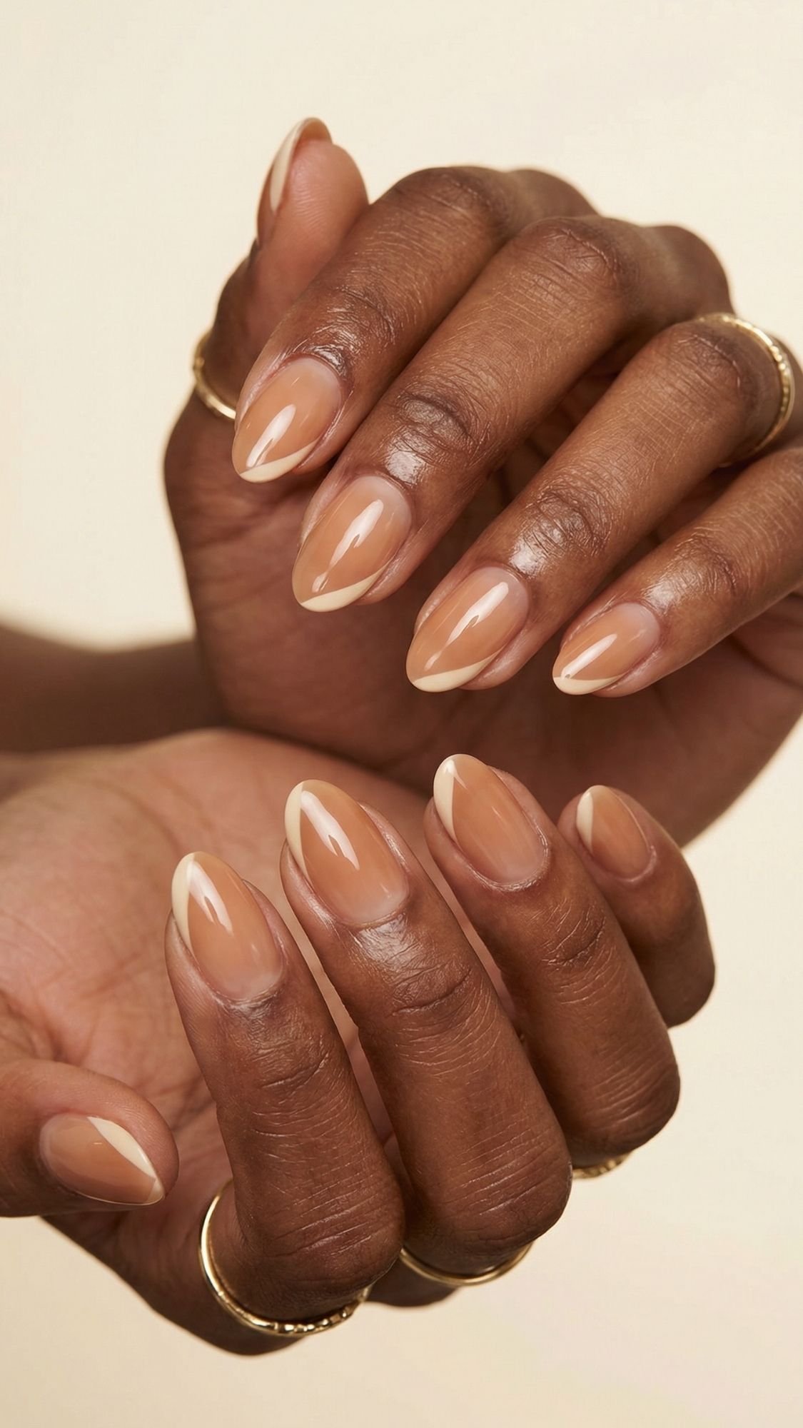 Close-up of hands with almond-shaped nails featuring a glossy nude French manicure—perfect for summer nails. The person is wearing thin gold rings on several fingers, set against a soft beige background.