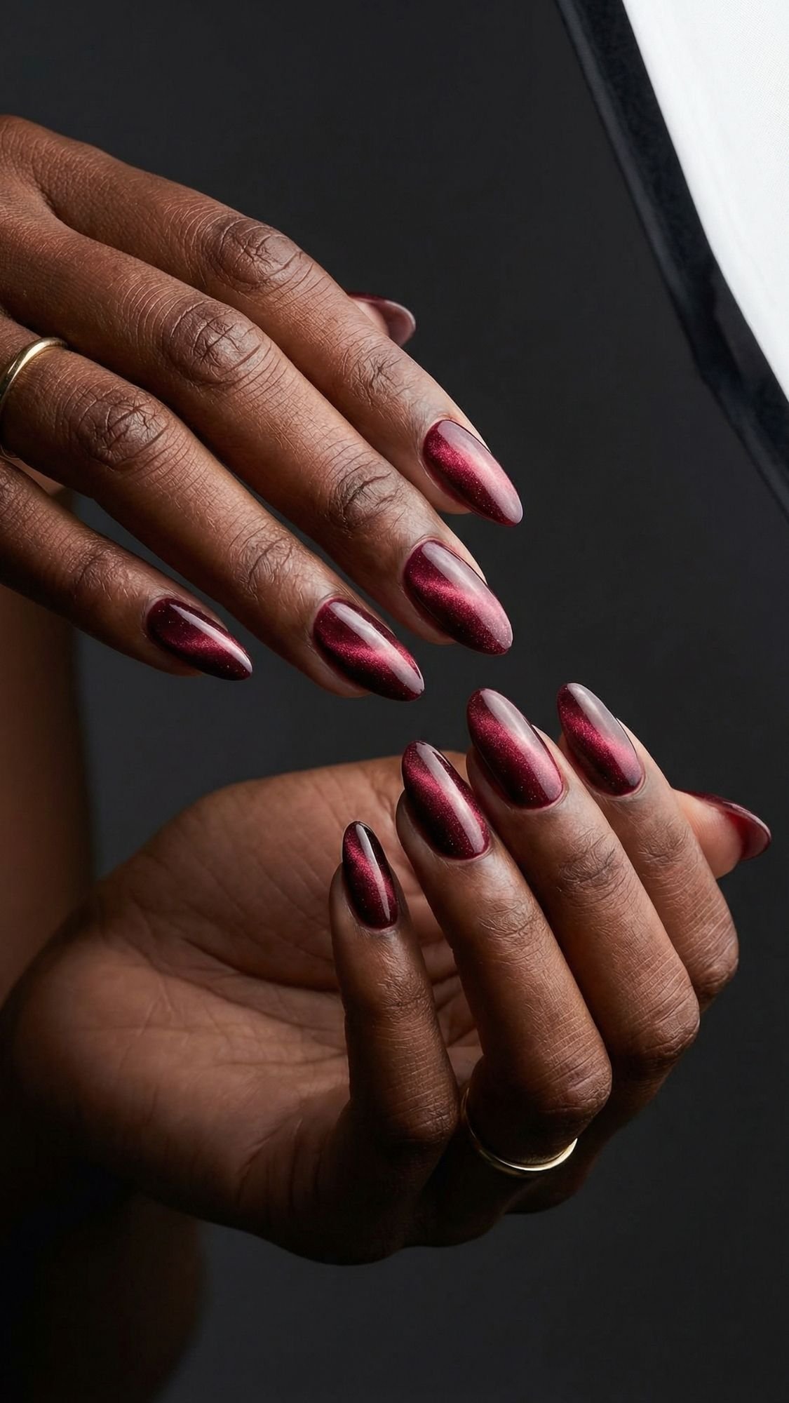 A close-up of hands with almond-shaped nails painted in glossy, metallic burgundy—a chic twist on red nails. Simple gold rings add elegance, making this look perfect for anyone seeking stylish seasonal nail designs against a dark backdrop.