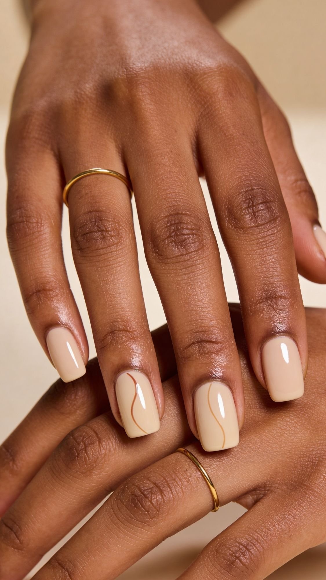 Two hands with medium brown skin and gold rings display manicured nails painted in a glossy nude shade, featuring a subtle, wavy tan line design on the ring fingernails—perfect inspiration for chic summer nails.