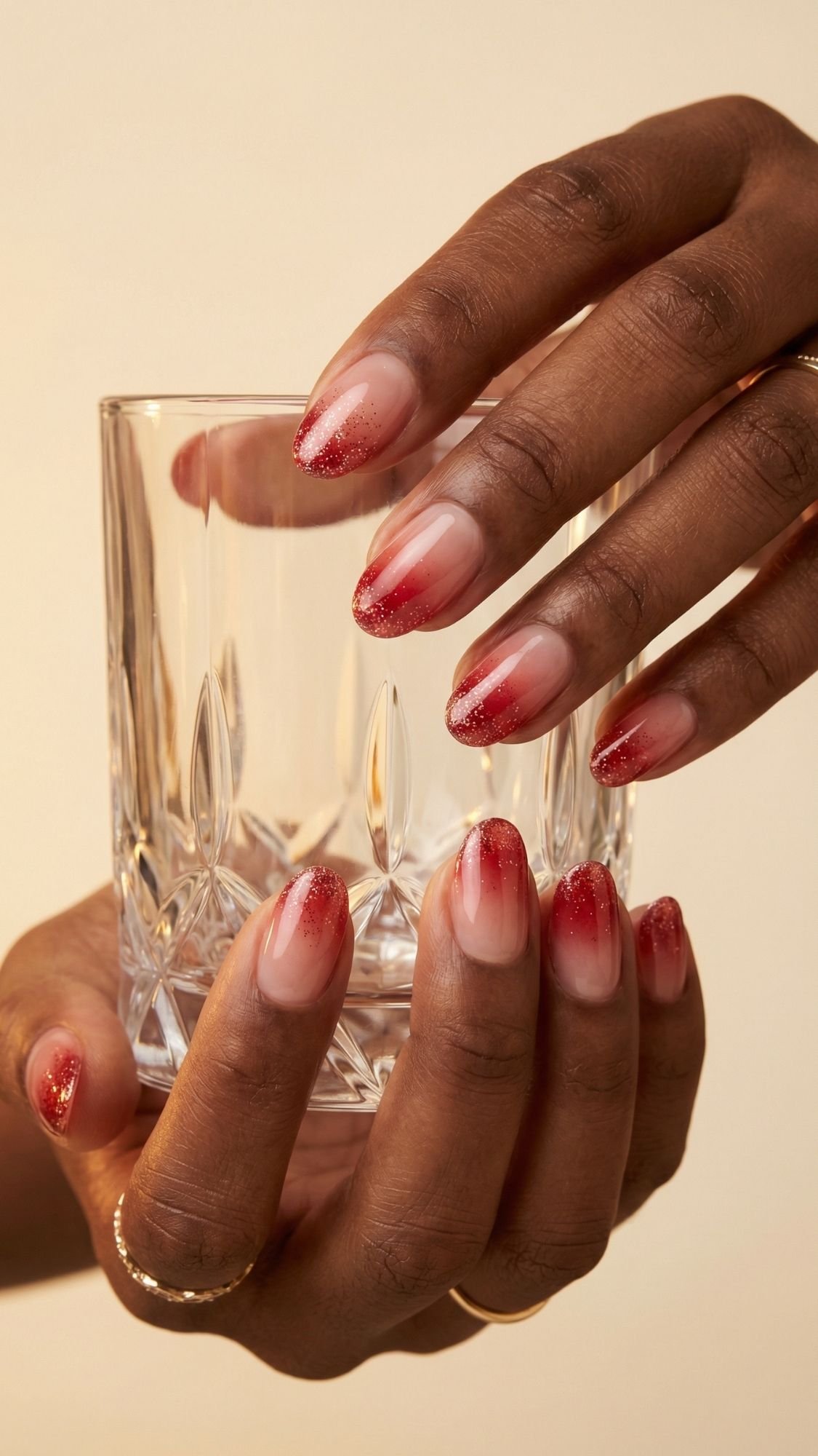 A hand with almond-shaped red nails featuring a glitter ombre manicure—perfect nail ideas for every season—holds a clear, faceted glass tumbler. The person wears thin gold rings on two fingers.