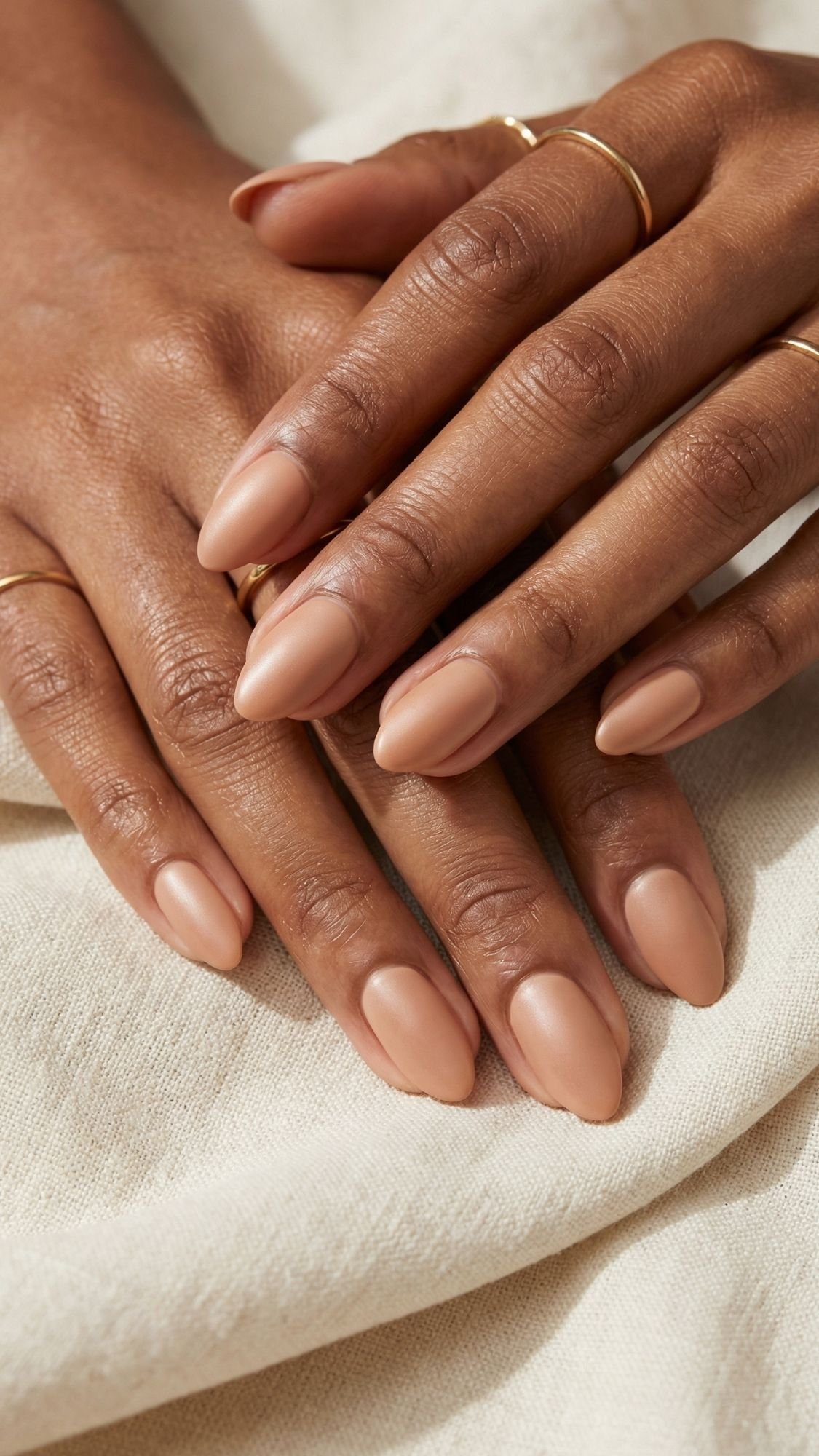 Close-up of hands with almond-shaped nails painted in a nude, glossy polish—perfect for Spring Nails. The person wears thin gold rings on several fingers, and their hands rest on soft, cream-colored fabric.