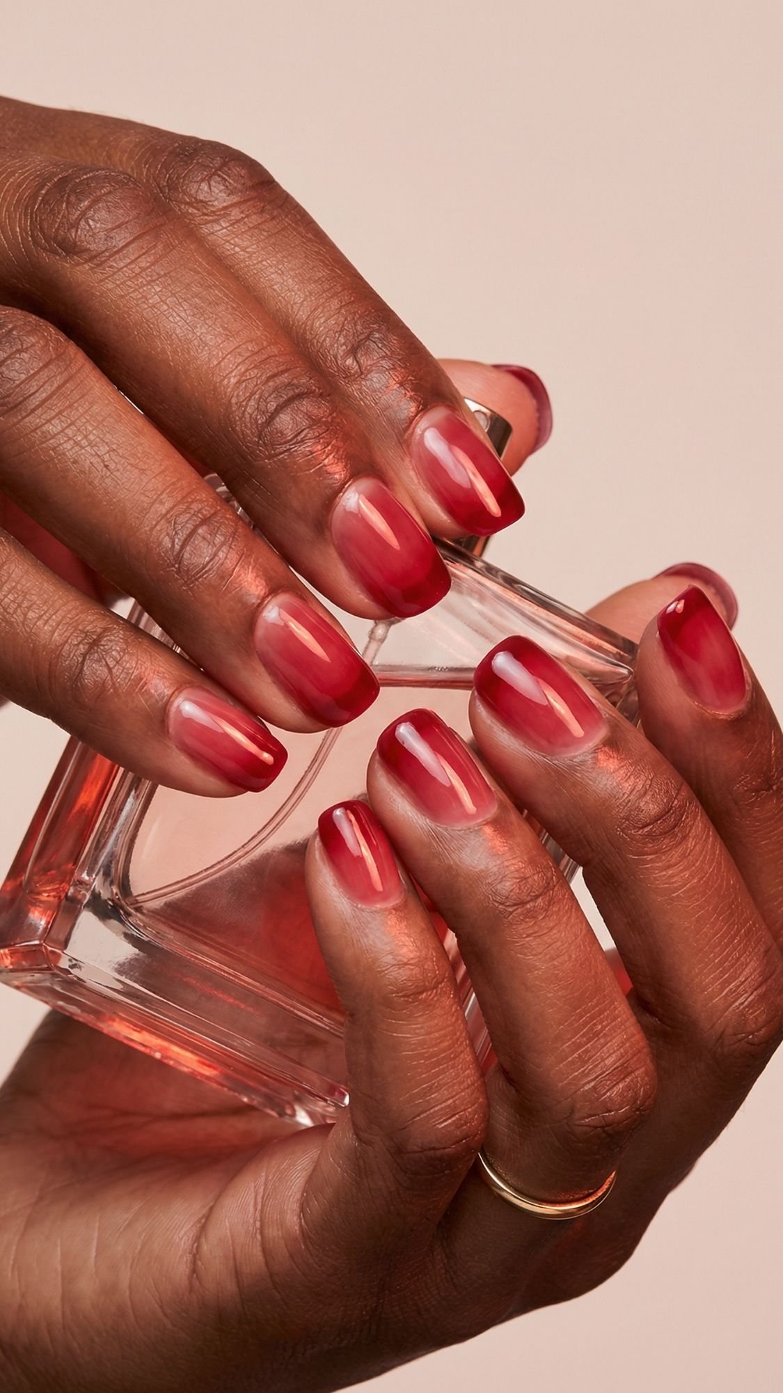 A close-up of hands with a gold ring on one finger, holding a glass perfume bottle. The glossy red ombré nails make a chic statement—an inspiring nail idea that works perfectly for every season.