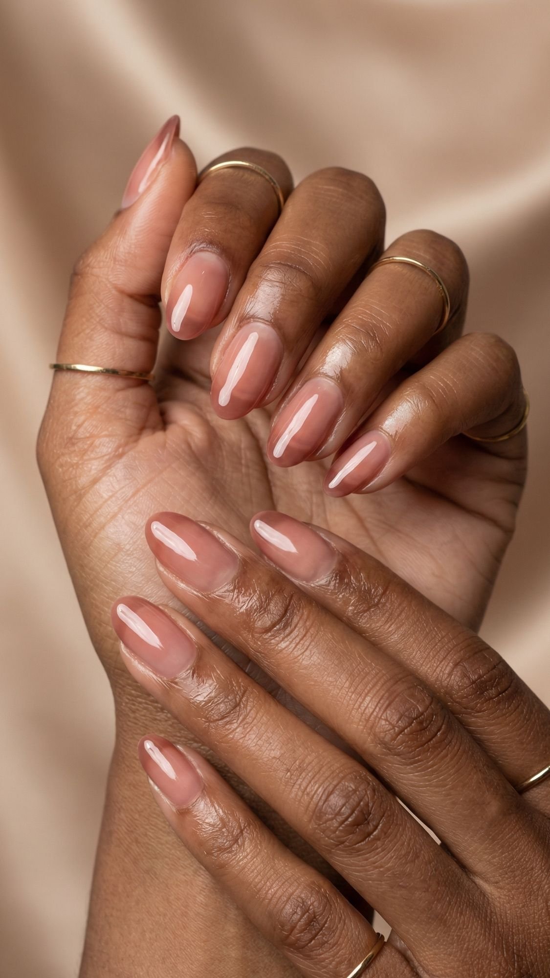 A close-up of hands with smooth, medium-brown skin, wearing thin gold rings and showcasing well-manicured summer nails painted with glossy nude-pink polish against a soft beige background.