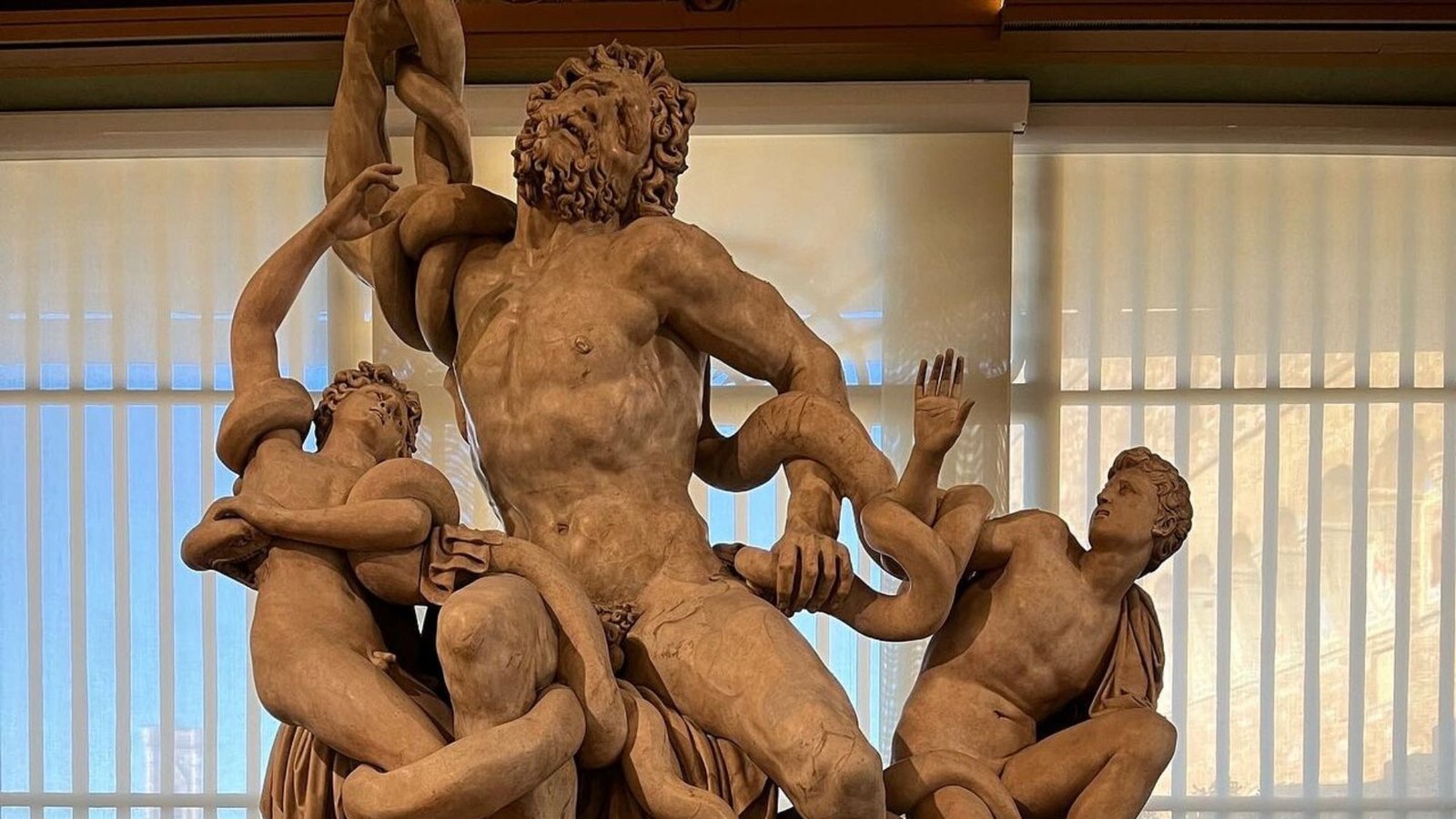 A classical sculpture from my first trip to Italy depicts three male figures entangled by snakes; the central bearded figure looks upward in agony as younger men struggle beside him. Vertical blinds cover a window in the background.