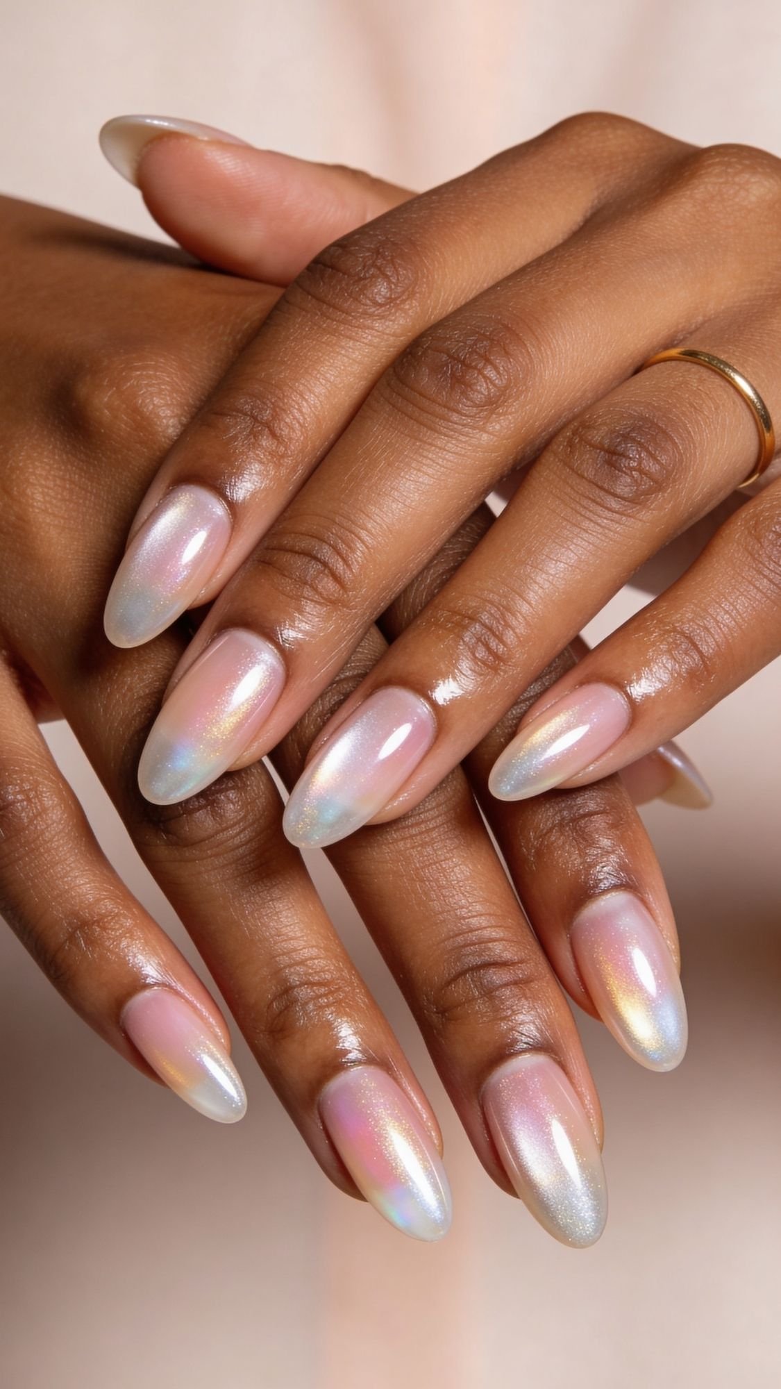 A close-up of two hands with almond-shaped, iridescent, pearly pink nails—an elegant example of easy nail designs you can try for simple nails at home. A simple gold band adds a touch of sophistication to the neatly manicured look.