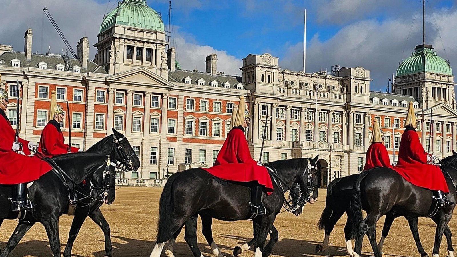 Horse guards in red uniforms ride black horses in front of a grand historic building with green domes—a must-see for your first trip London and one of the top London things to do under a partly cloudy sky.
