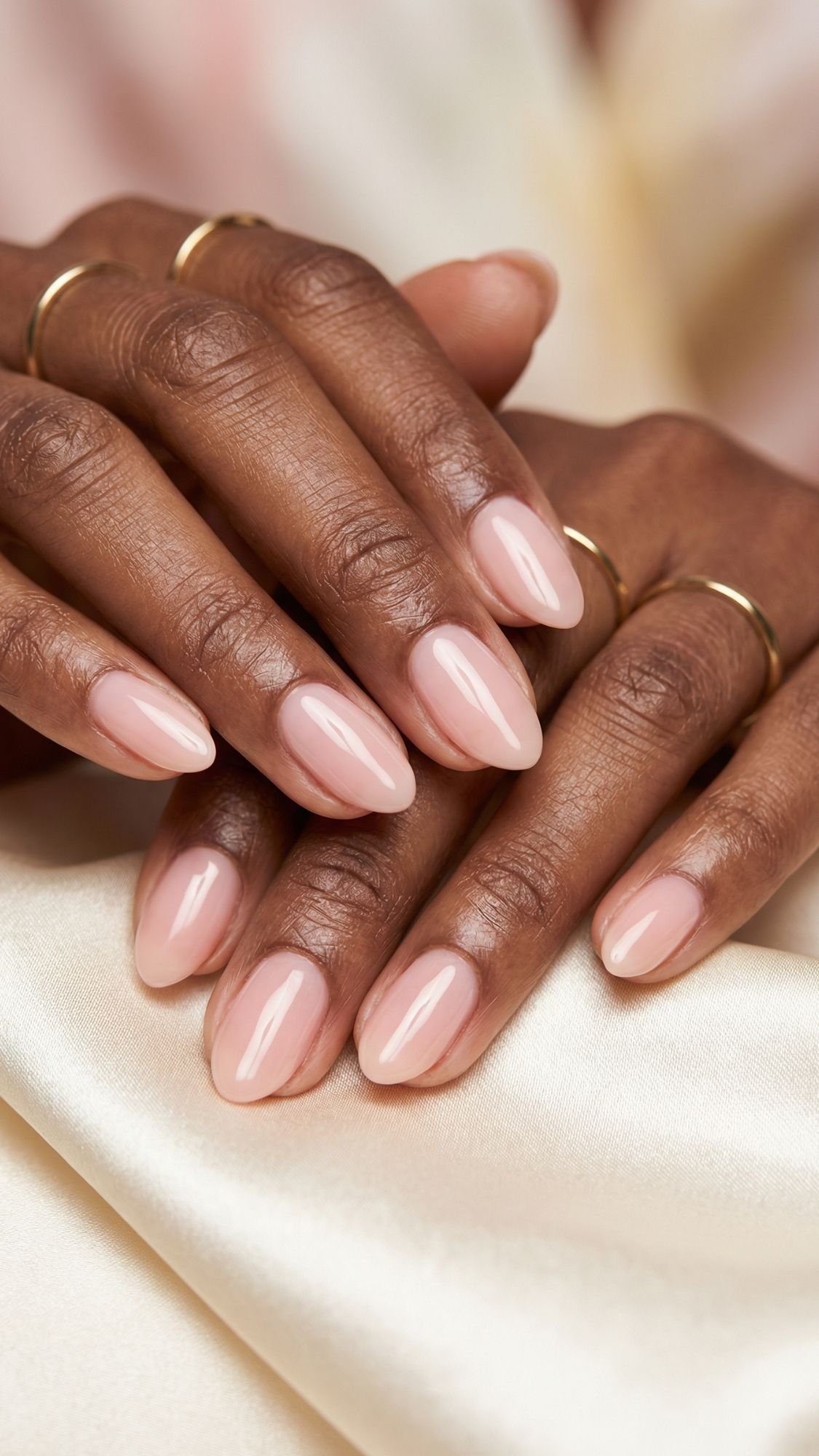 Close-up of well-manicured hands with almond-shaped nails painted in a glossy nude pink shade, perfect for summer nails, resting on cream satin fabric. The person is wearing simple gold rings on several fingers.