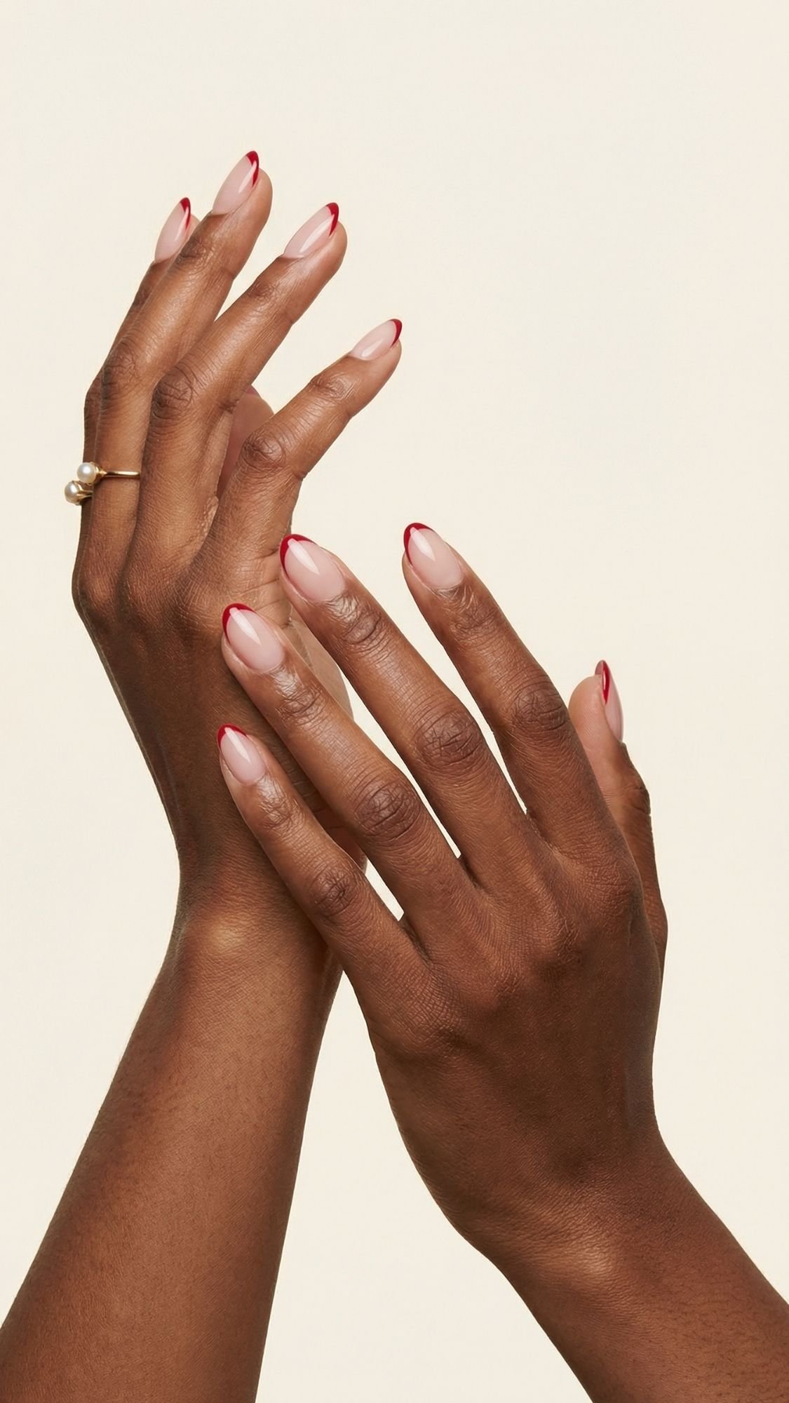 Two hands with medium brown skin are held gracefully against a light background. The almond-shaped nails feature a glossy nude base and thin red tips—an elegant take on red nail designs. One hand wears a gold ring with a single pearl.