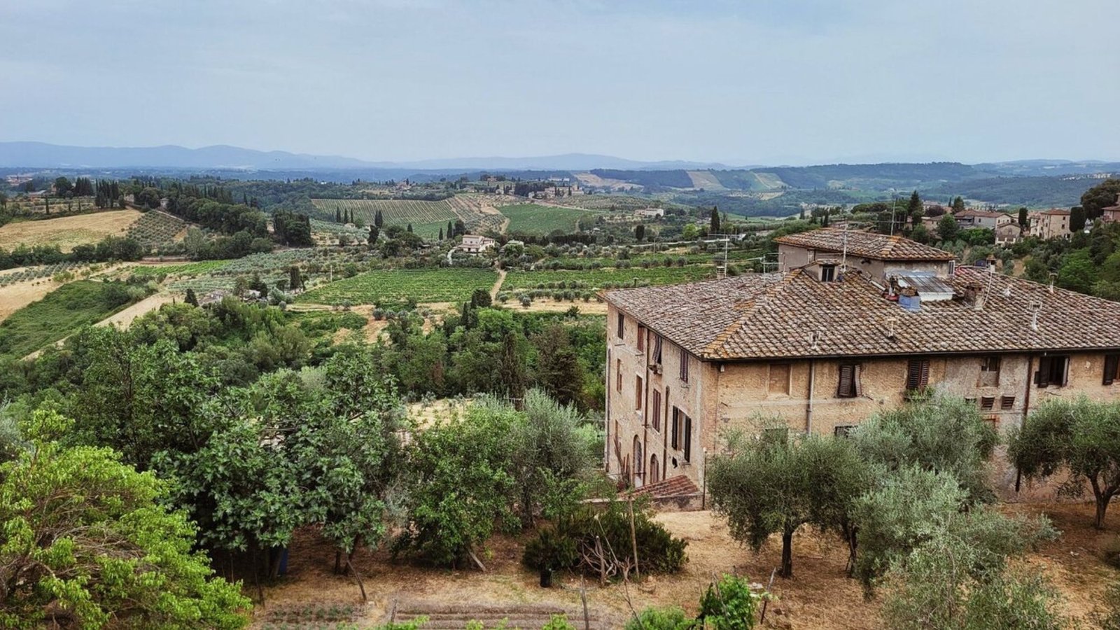 A rustic building with a tiled roof sits among trees and olive groves, overlooking green rolling hills—a perfect scene for your Italy bucket list or an unforgettable first trip to Italy.