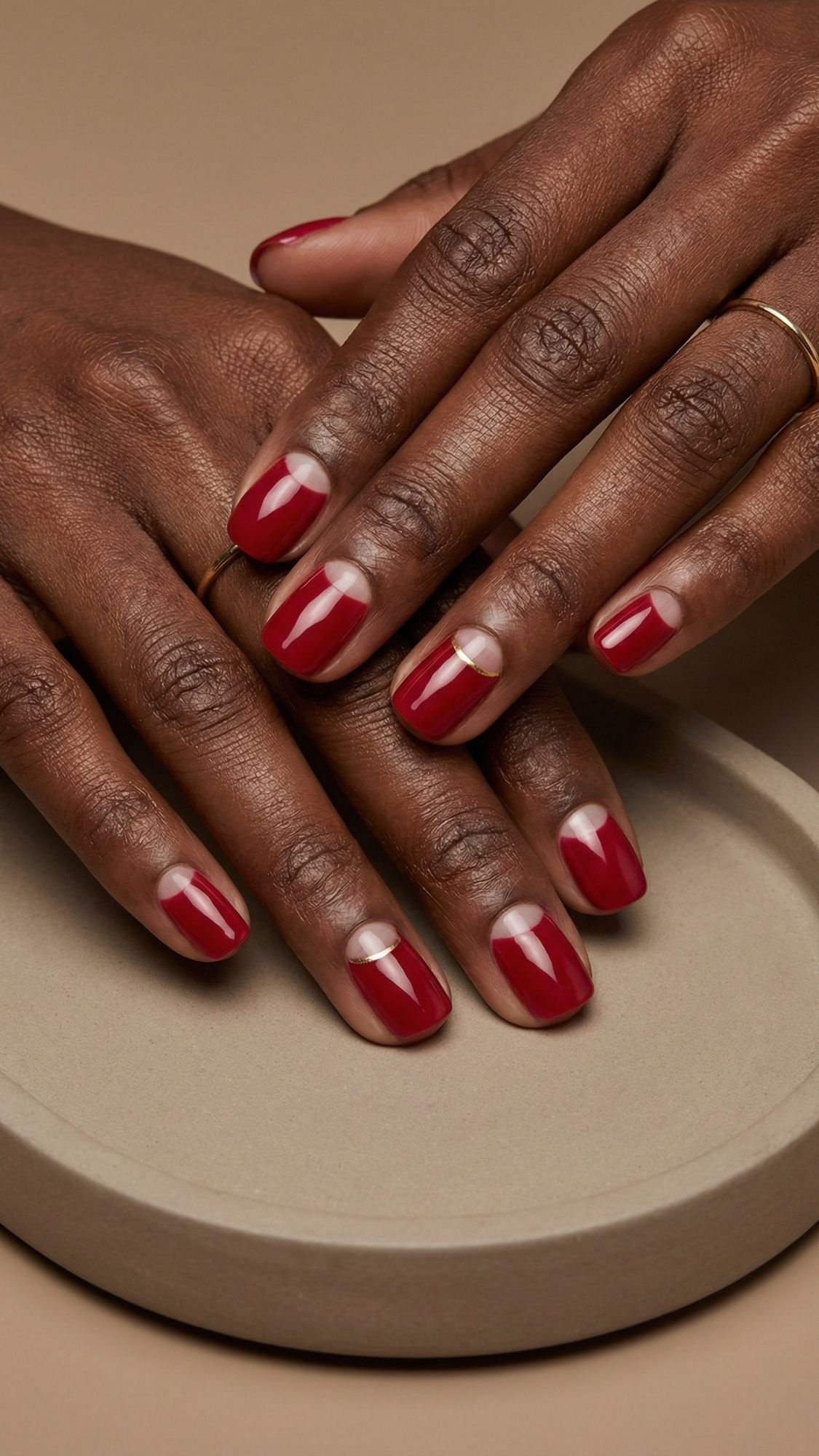Close-up of hands with a minimalist manicure featuring deep red nails and bare half-moon shapes at the base, resting on a round, neutral surface. Perfect inspiration for red nails for every season, with a thin gold ring adding elegance.