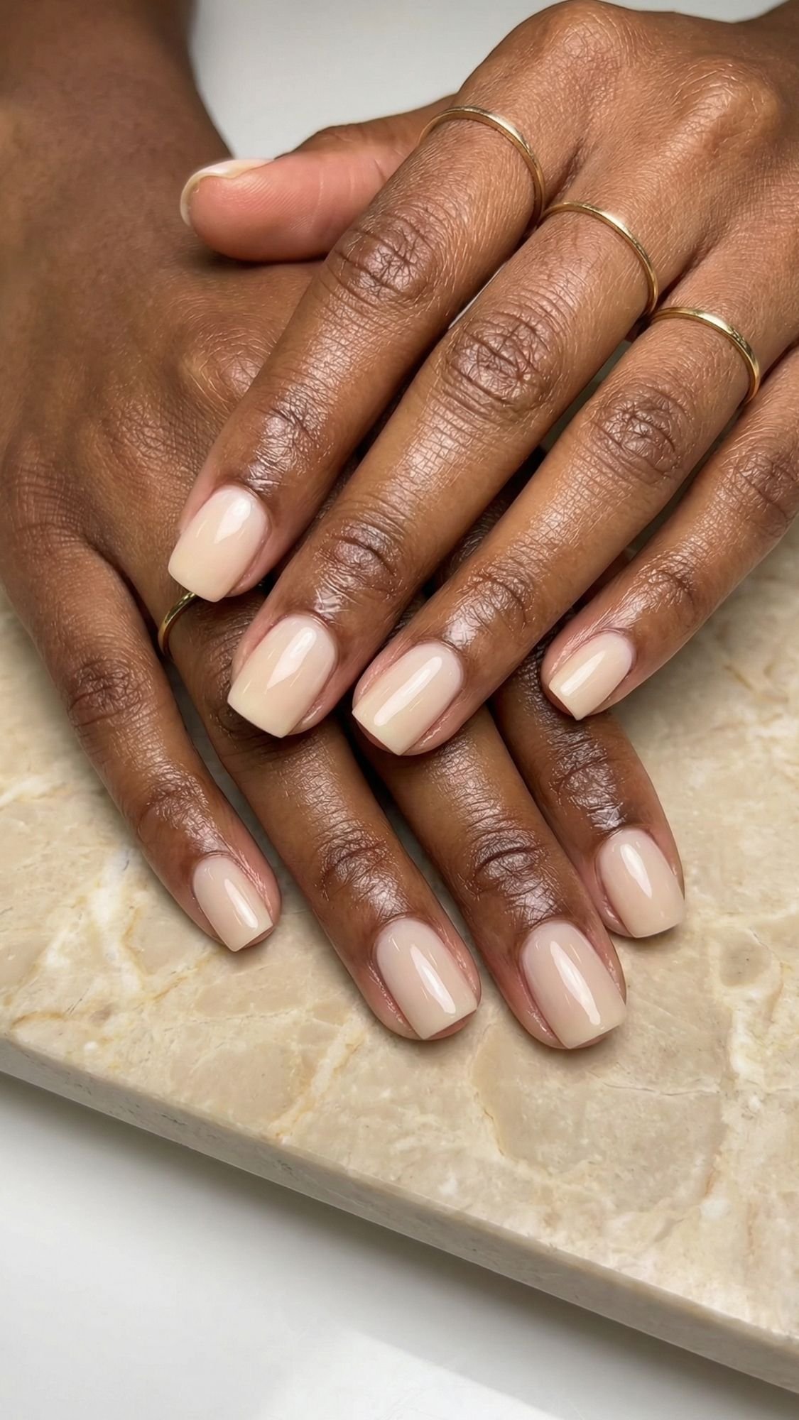Two hands with neatly manicured summer nails painted in a glossy nude color rest on a beige marble surface. The person is wearing thin gold rings on several fingers—perfect inspiration for chic nude nail ideas.
