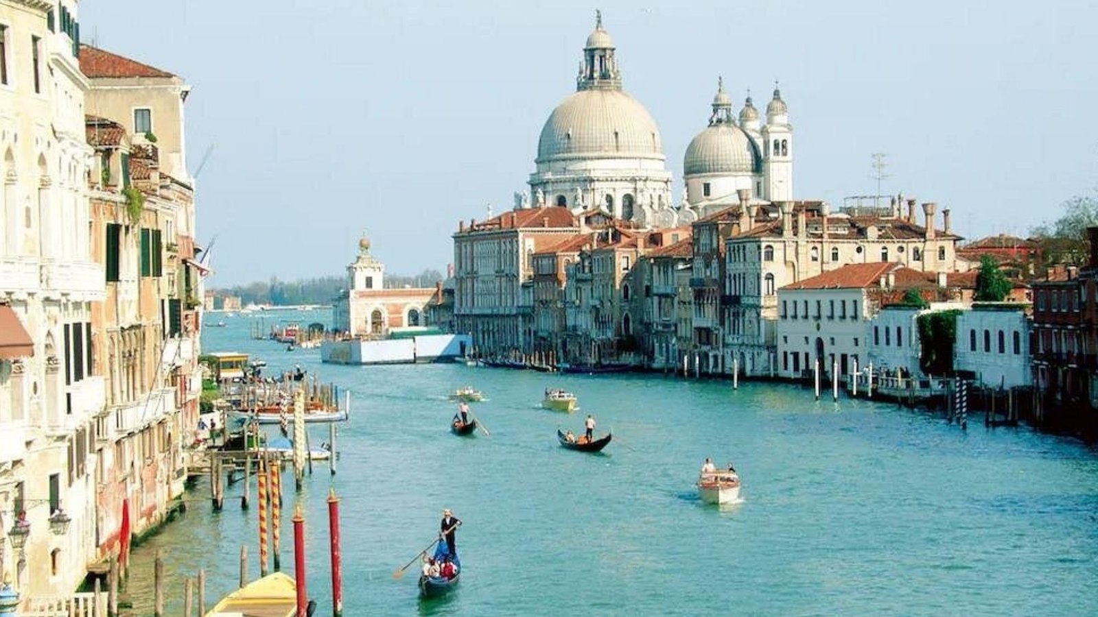 A wide canal in Venice, Italy, lined with historic buildings and gondolas on the water—an essential stop for any Italy Bucket List. The domed Santa Maria della Salute church stands prominently in the background under a clear sky.